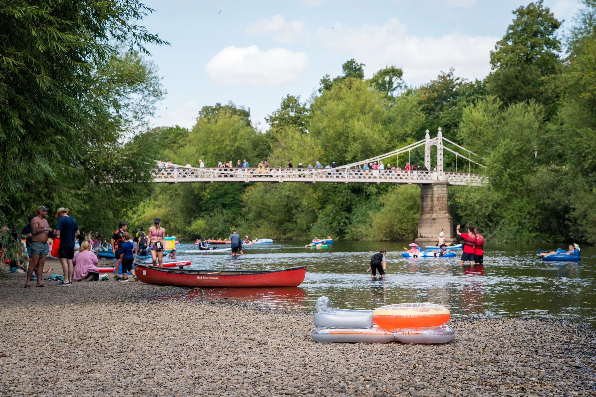 People enjoying a river day with canoes, kayaks, and inflatable floats. A bridge crosses over the river, surrounded by lush green trees. Some individuals are on the rocky shore while others are paddling or floating in the water.