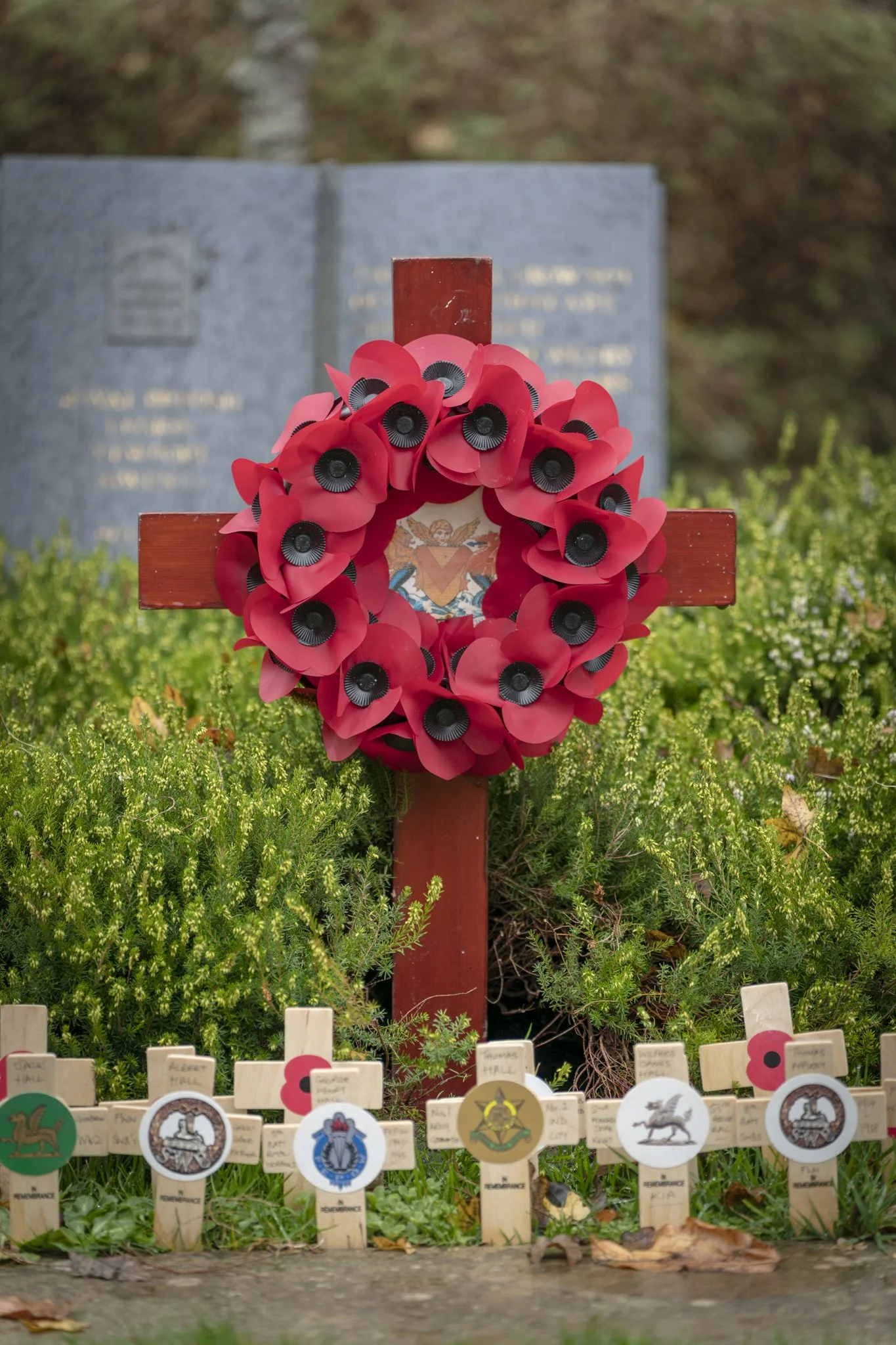 A red cross adorned with red paper poppies on a pink background stands above small wooden memorial plaques in front of a larger grey headstone.