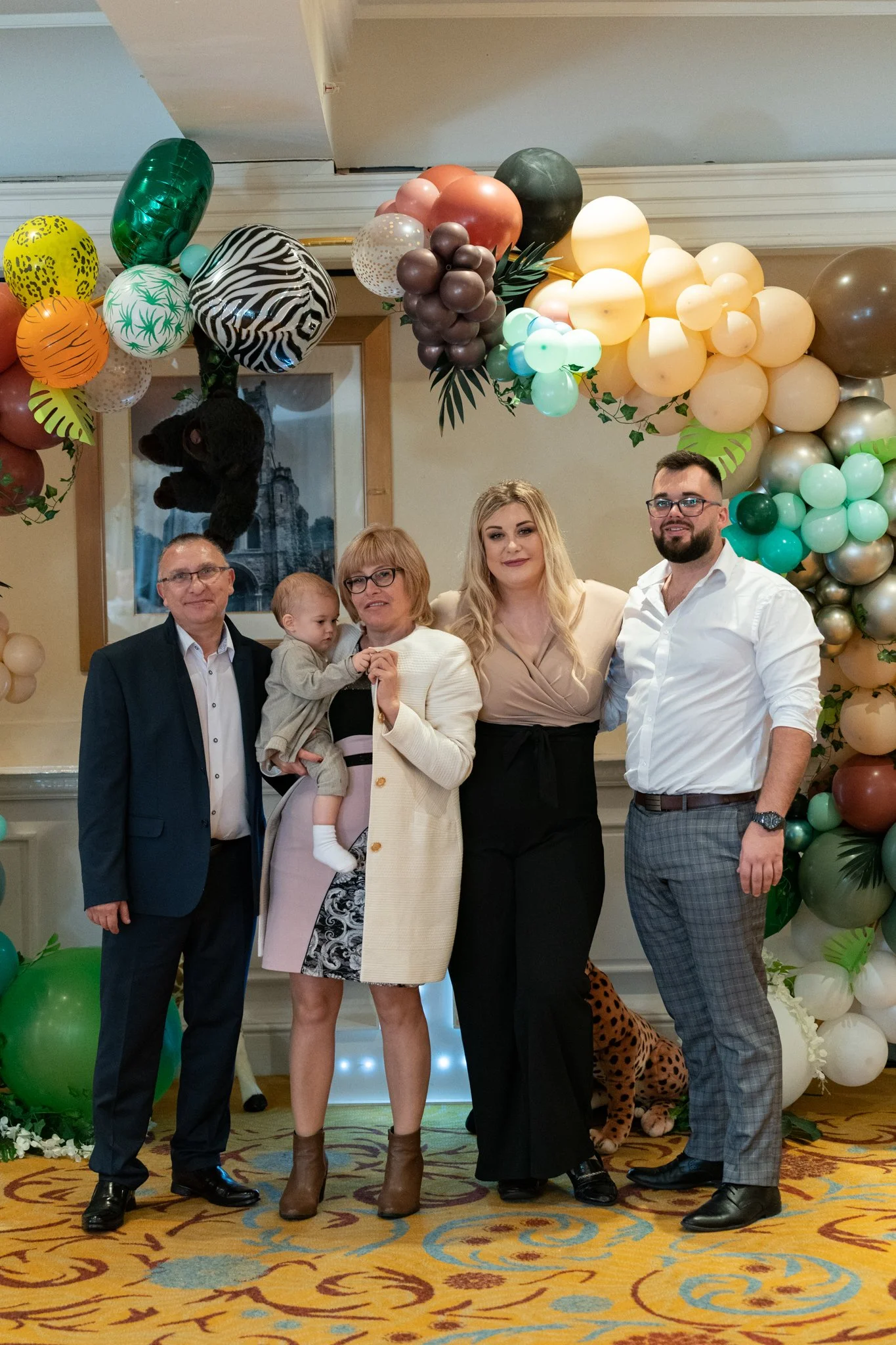 Group of five adults and one child standing indoors, posing for a photo, with balloon decorations in natural colors and animal print design in the background.