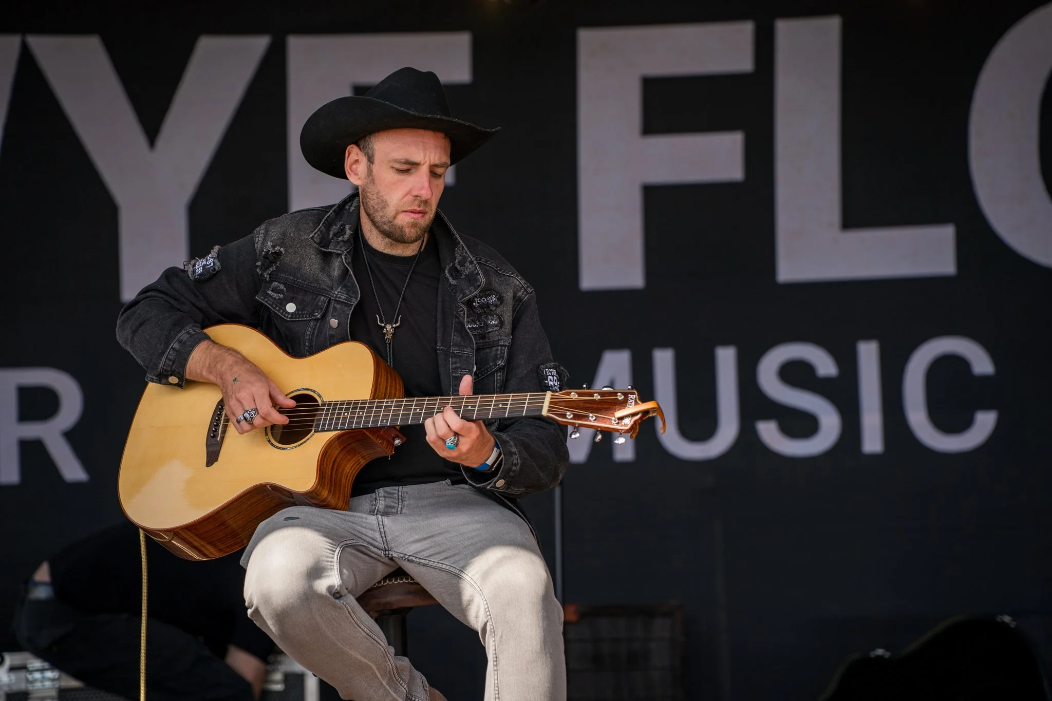Man wearing a black cowboy hat, distressed black denim jacket, and beige pants, playing an acoustic guitar on stage.