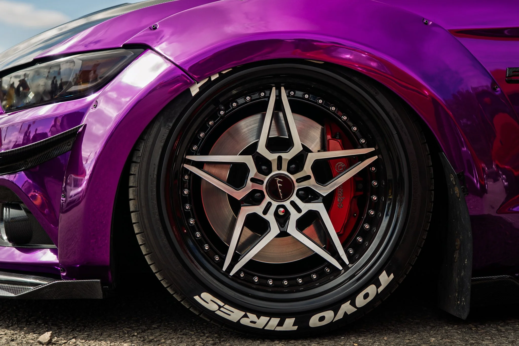 Close-up of a purple sports car's front wheel, featuring a black and silver rim with the Mercedes-Benz logo at the center and red brake calipers behind the rim. The car's body has a shiny, reflective purple paint.
