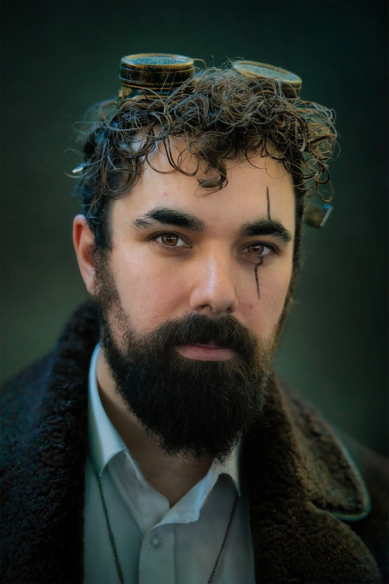 Close-up of a man with dark curly hair and a beard, wearing a white shirt and a brown coat. He has a black line drawn vertically down his face, crossing his right eye, and a line on his forehead. 