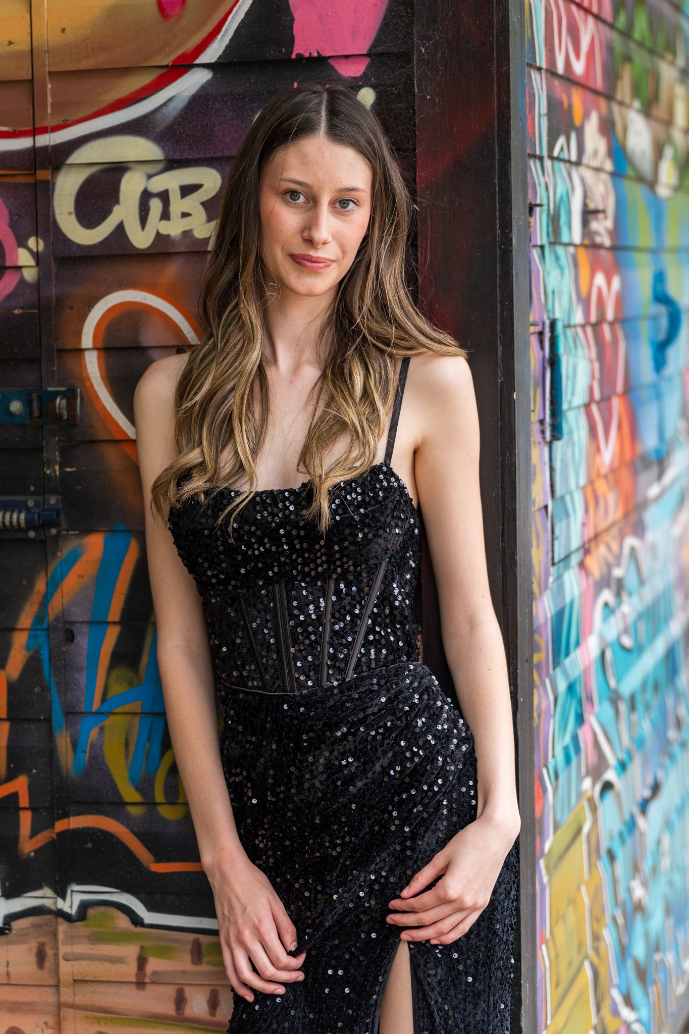 A young woman with long, wavy brown hair and fair skin wearing a black, sequined, strapless dress with a slit, standing in front of a colorful graffiti wall.