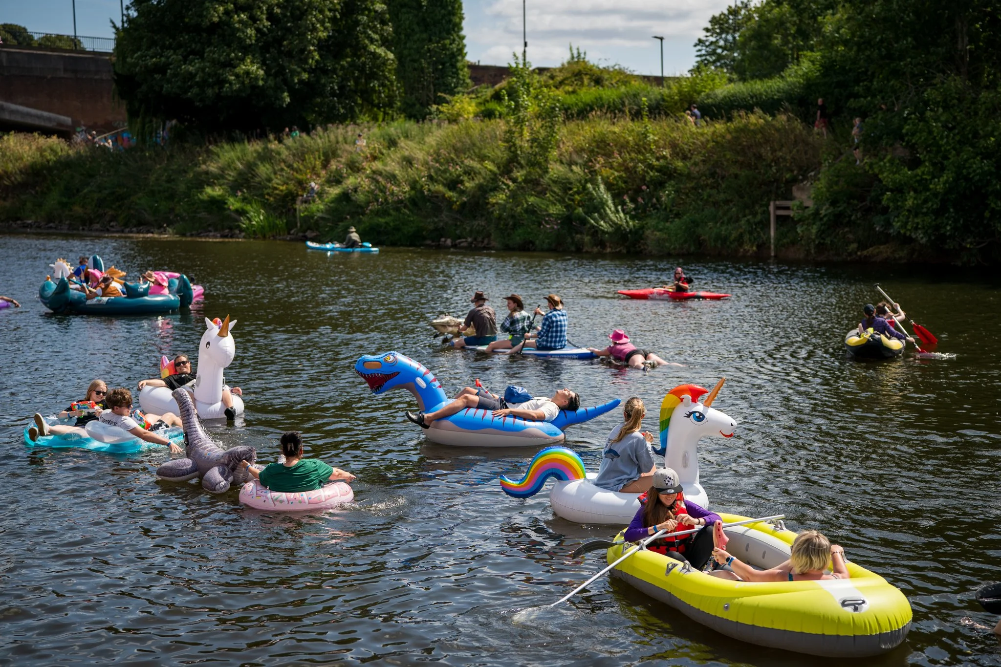 People floating on a river in inflatable boats and pool floats shaped like unicorns, dinosaurs, and rainbows, with a grassy bank and trees in the background.