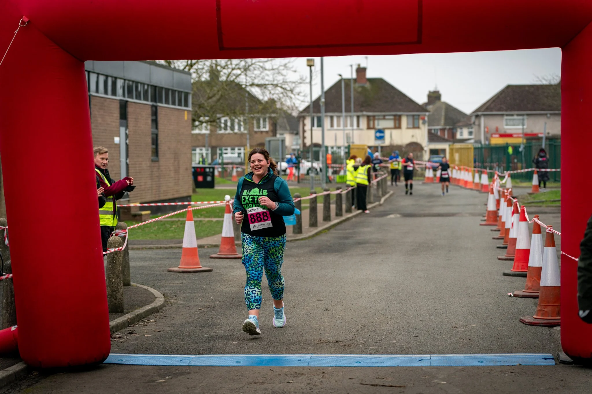A woman running in a marathon crossing the finish line under a red inflatable arch, with spectators and volunteers along the road in a residential area.