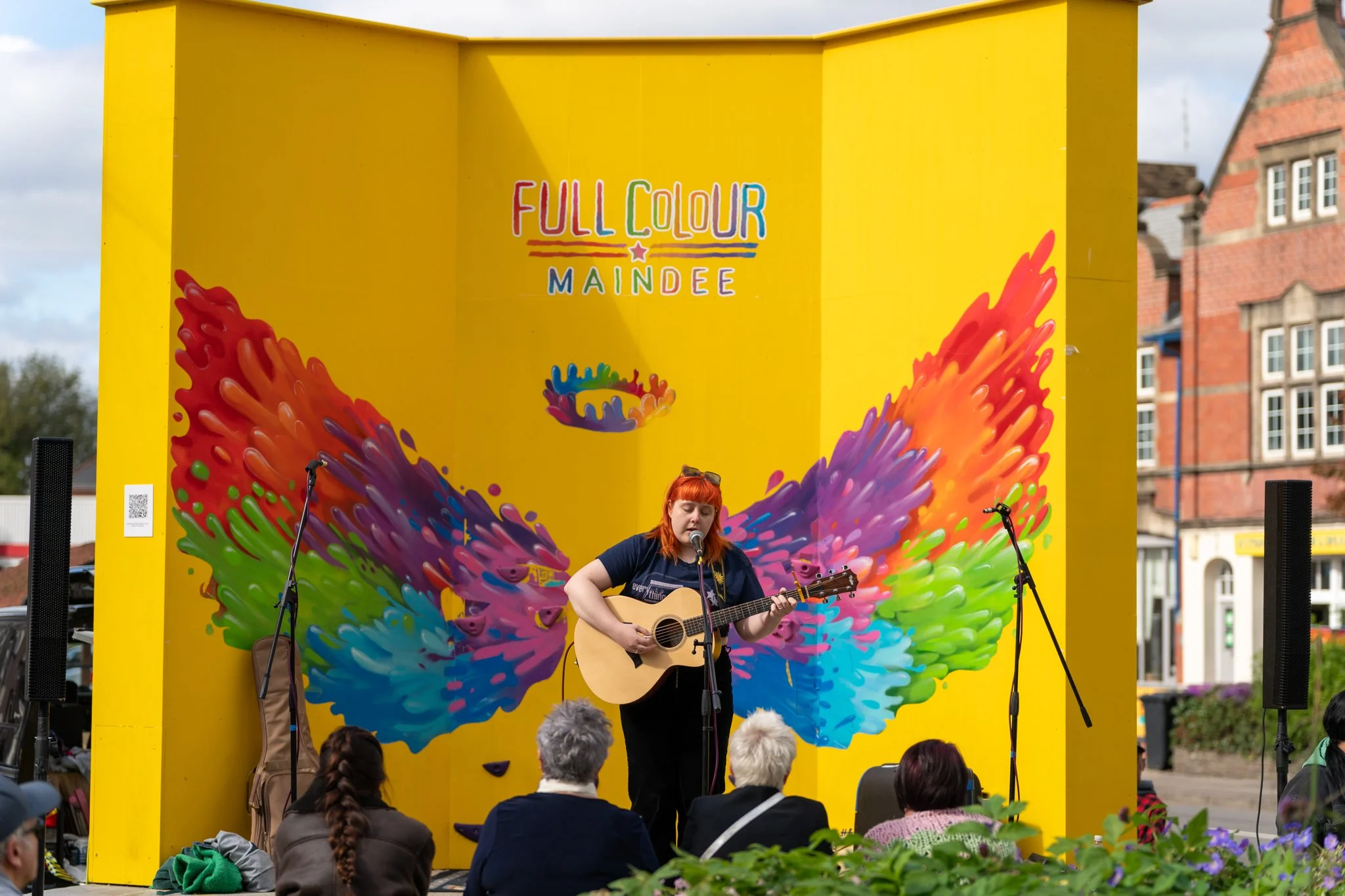 A woman with red hair singing and playing guitar on a stage with a bright yellow background, decorated with colorful splash and swirl designs, at an outdoor event in front of an audience.
