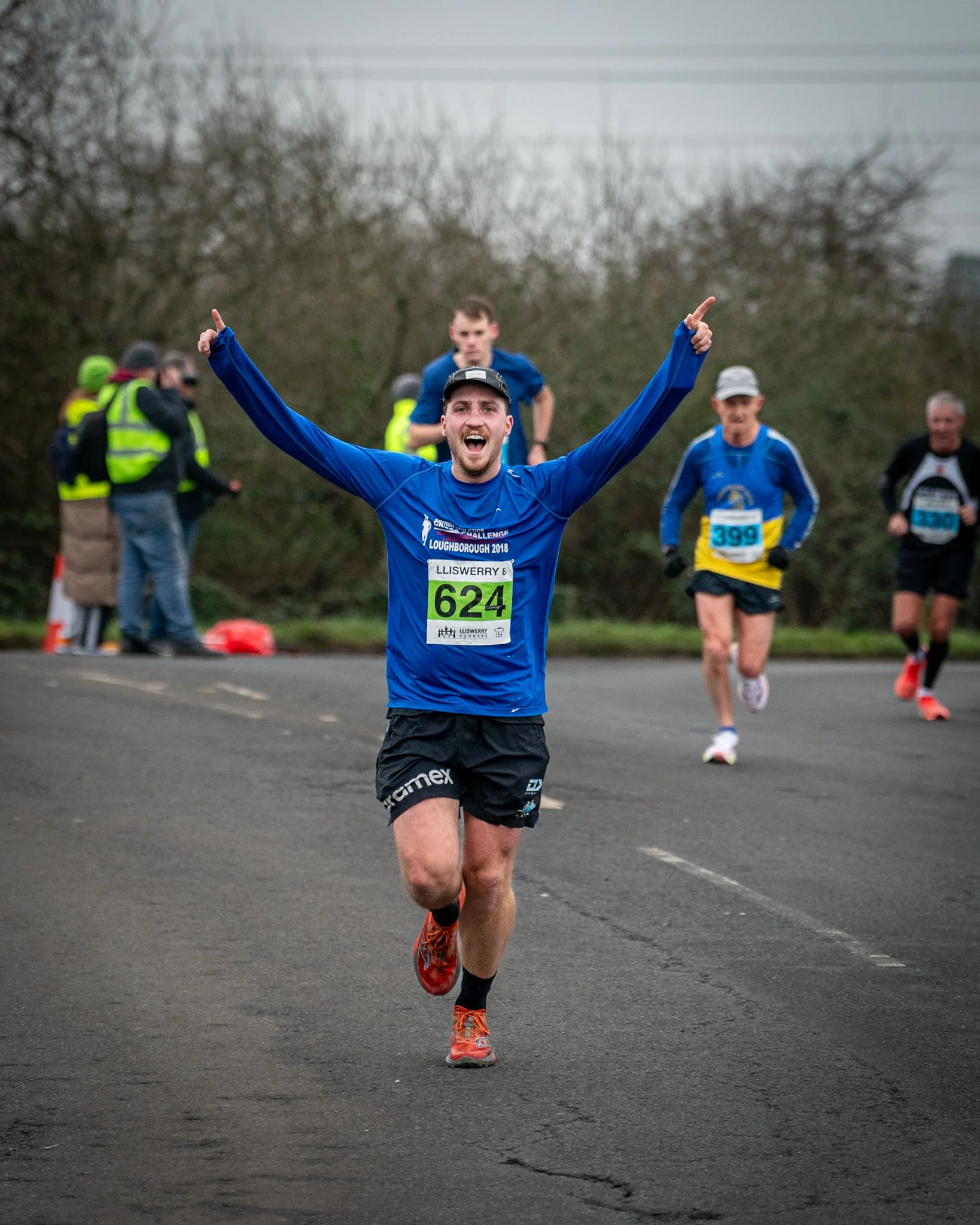 Male runner with arms raised celebrating during a race, other runners and event volunteers in the background, on an asphalt track outdoors.