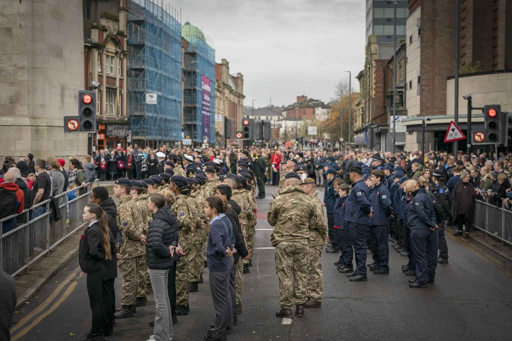 A large crowd gathered on a city street, with many people in military and police uniforms standing behind barricades. Traffic lights are visible, and buildings with scaffolding are in the background.