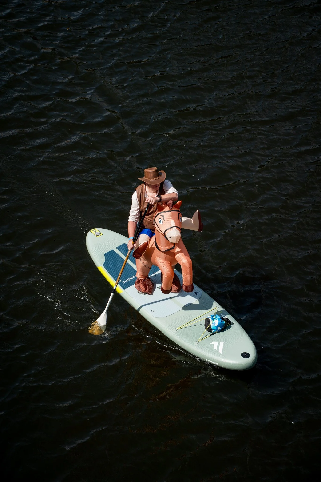 Person paddleboarding on a body of water, wearing a cowboy hat and a beige vest, with an inflatable horse attached to the front of the paddleboard, and a small bag and paddles secured on the board.