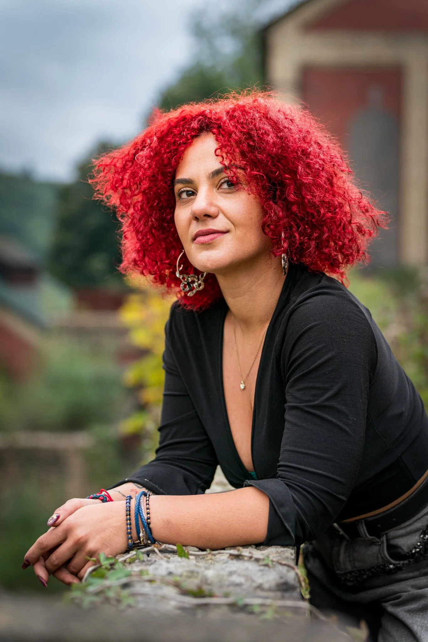 A woman with bright red, curly hair wearing a black top, earrings, and multiple bracelets poses outdoors with her arms resting on a stone surface, with a blurred background of trees and buildings.