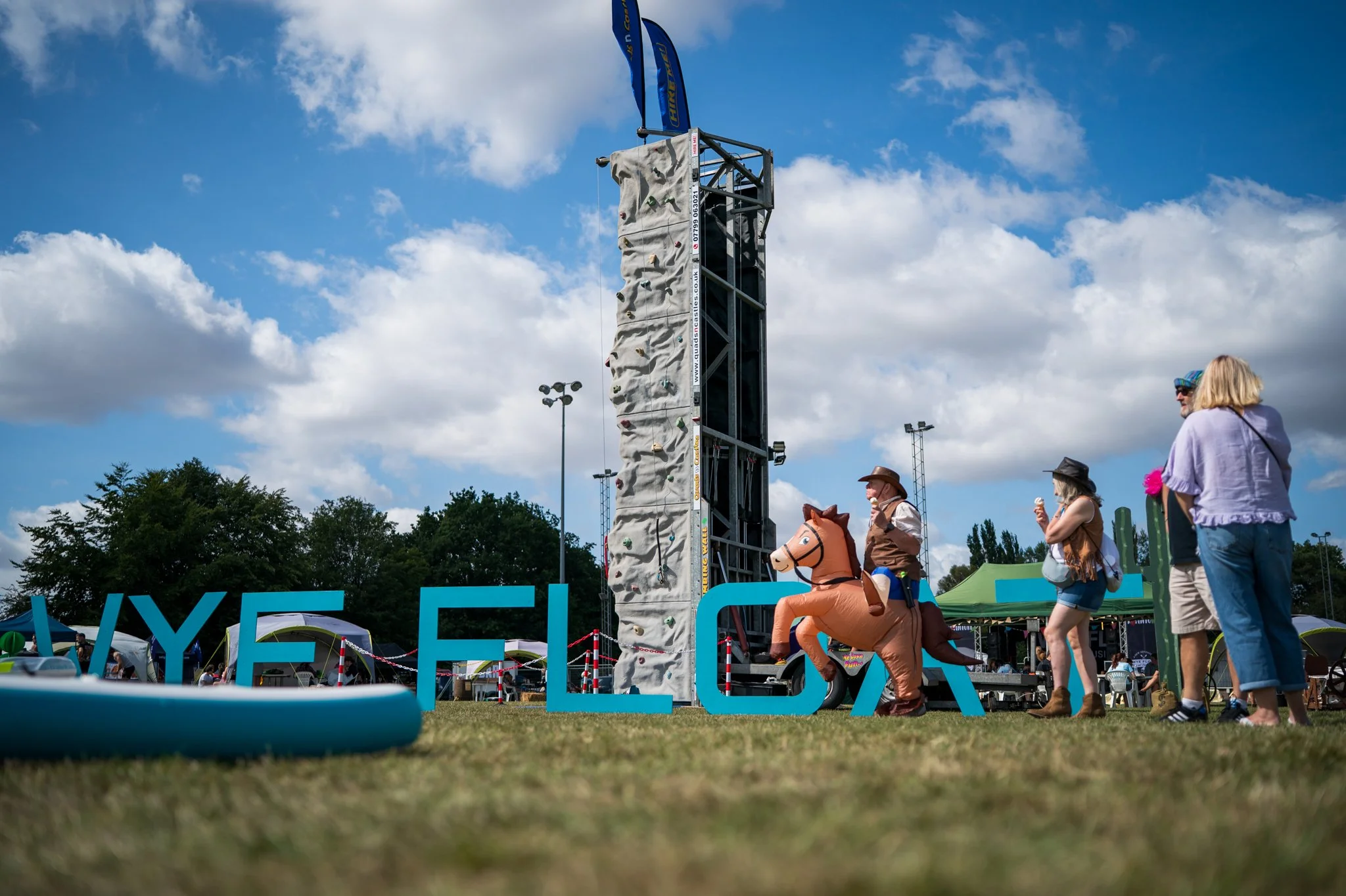 An outdoor event with a climbing wall and a person riding a horse costume in front of large blue letters spelling 'WYE FLOAT'. Several people are standing nearby, some wearing hats and sunglasses, under a partly cloudy sky.