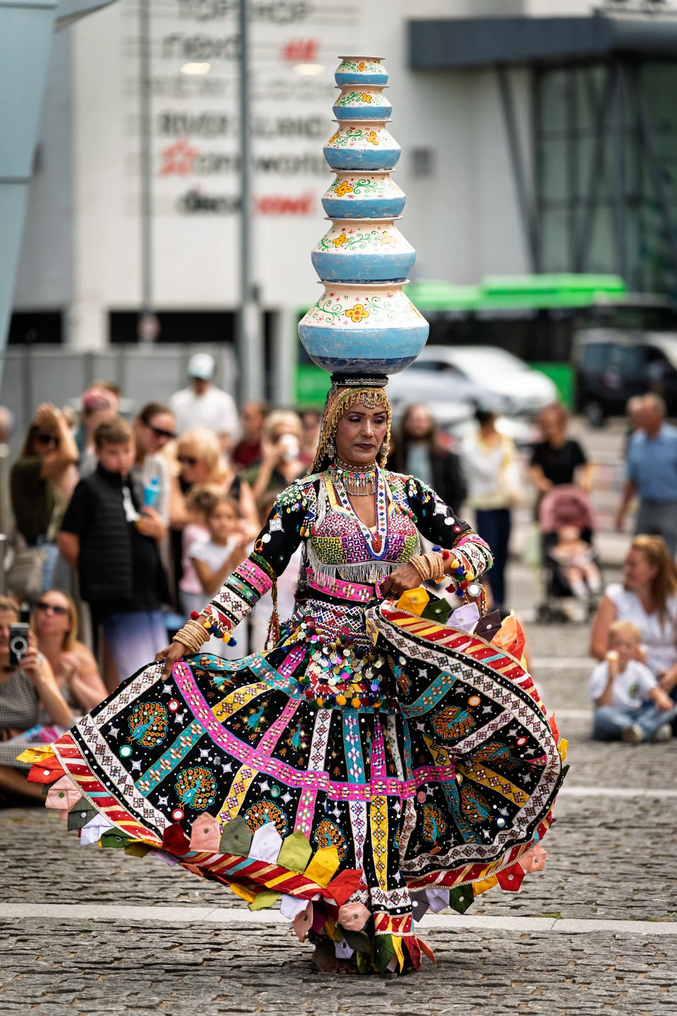 A woman dressed in a brightly colored, embroidered traditional costume spinning in a public square during a cultural festival. She balances a tall, stacked arrangement of painted bowls on her head. Spectators watching her in the background.