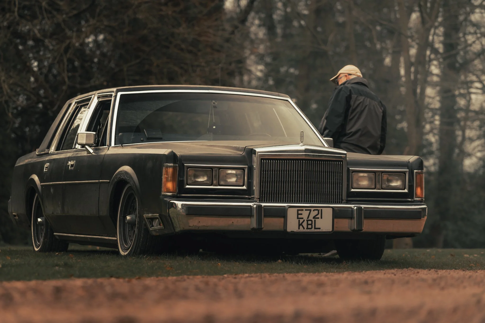 A man with glasses and a beige cap standing beside a black vintage car with a boxy design in an outdoor setting with trees in the background.