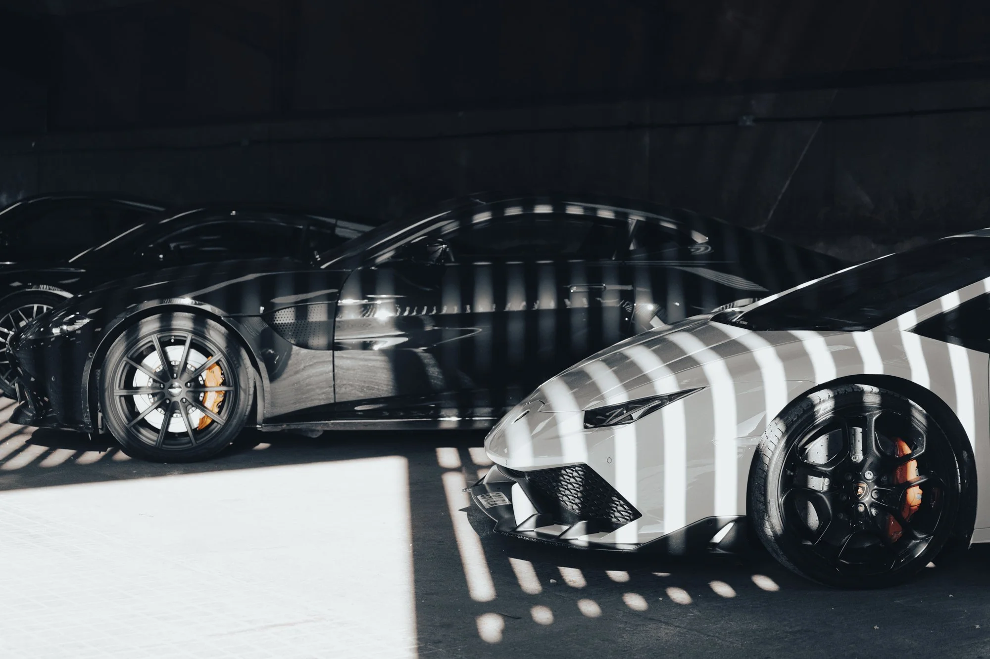 Multiple luxury sports cars, including a black car and a white car, parked in a dark garage with striped shadow patterns on the ground.