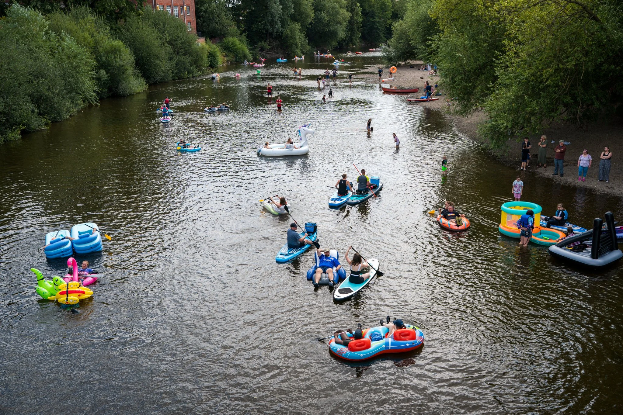 People enjoying a sunny day on a river with various floatation devices, including inflatable swans, unicorns, and kayaks, surrounded by lush green trees and a few spectators on the riverbank.
