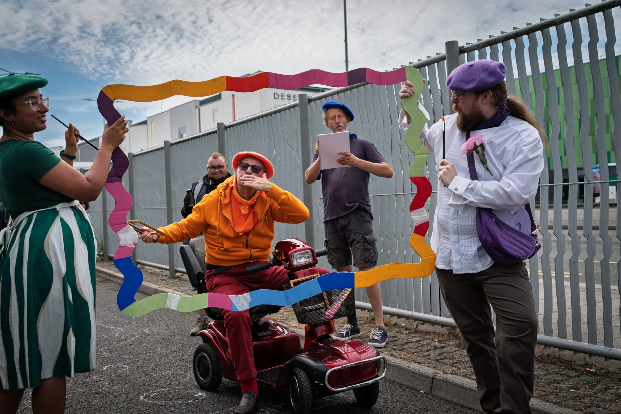 Group of diverse people participating in an outdoor event, some wearing colorful berets, standing around a red mobility scooter with a man in orange sitting on it, holding a rainbow-colored paper chain, with a gray fence and cloudy sky in the backgro