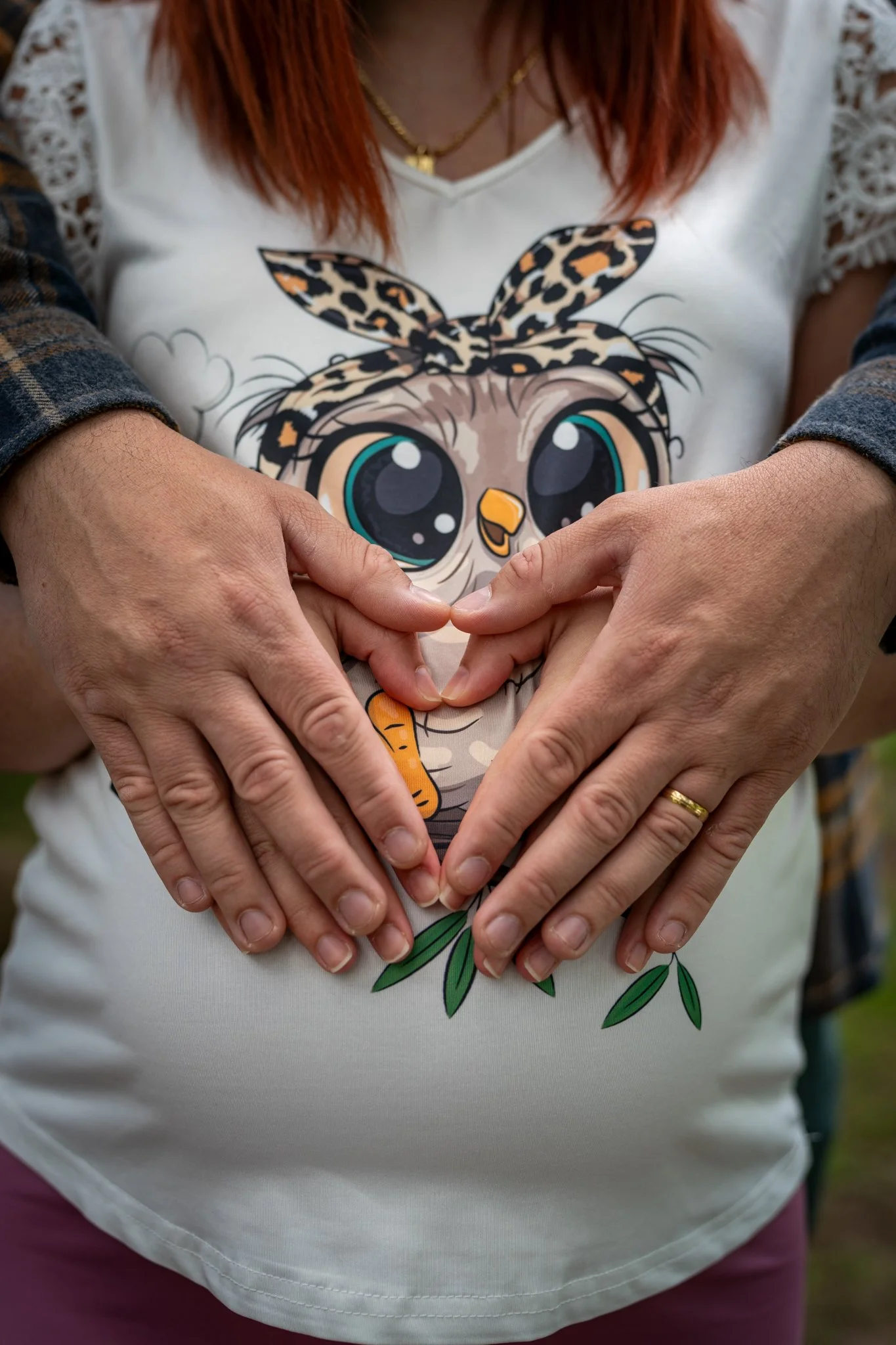 A person is making a heart shape with their hands, which are in front of their stomach. They are wearing a white shirt with a cartoon owl featuring large eyes, a beak, and a headband with leopard print. Maternity shoot.