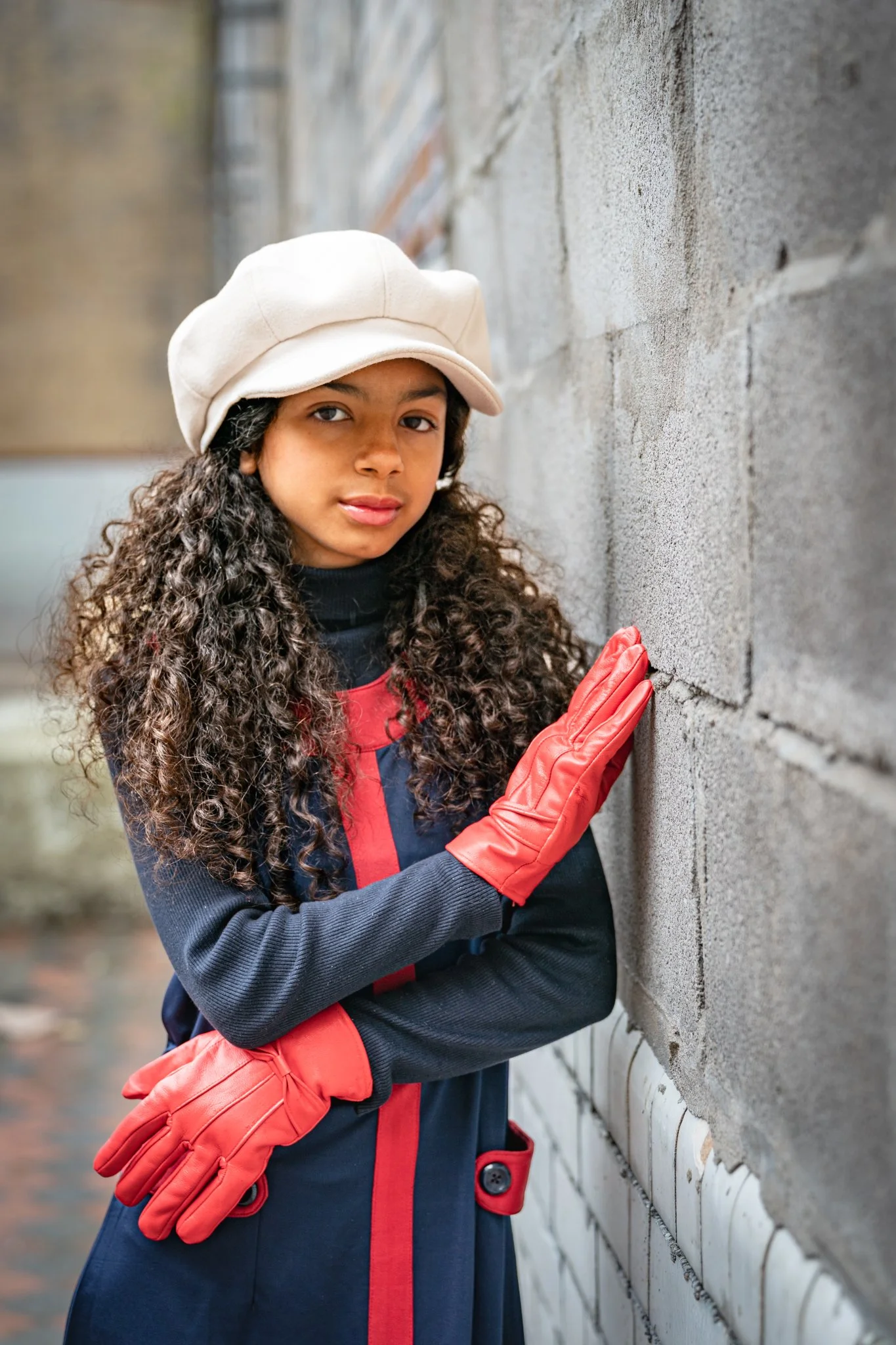 A young girl with curly hair wearing a beige cap, red gloves, and a dark-colored jacket with red accents, leaning against a concrete wall with her hand on it.
