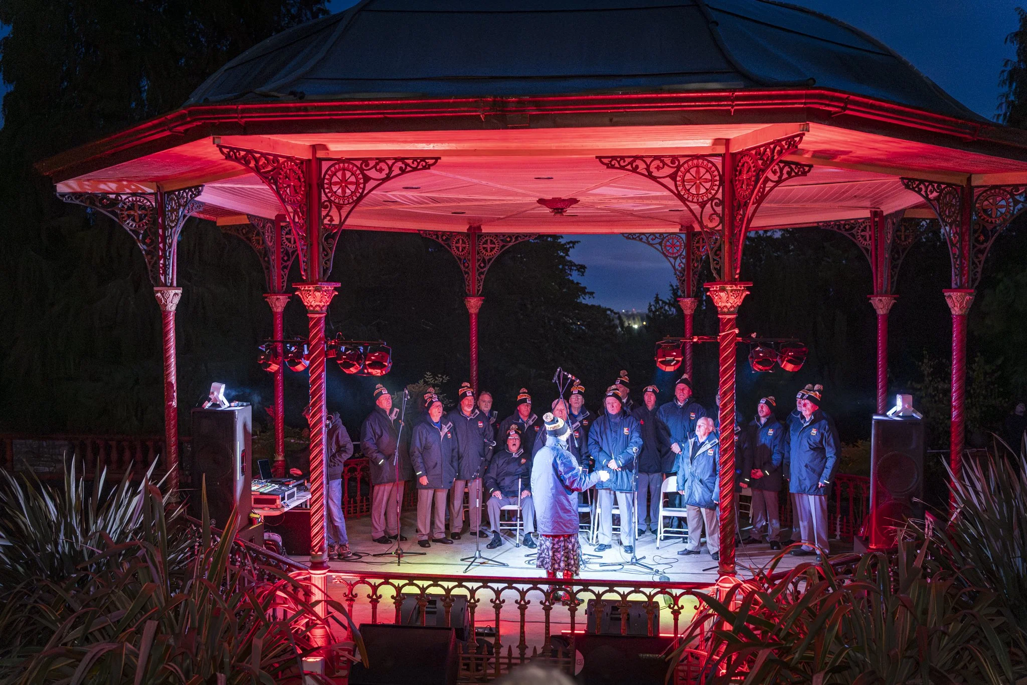 People performing in a choir on an outdoor stage at night, under a decorative red lit gazebo, with trees in the background.