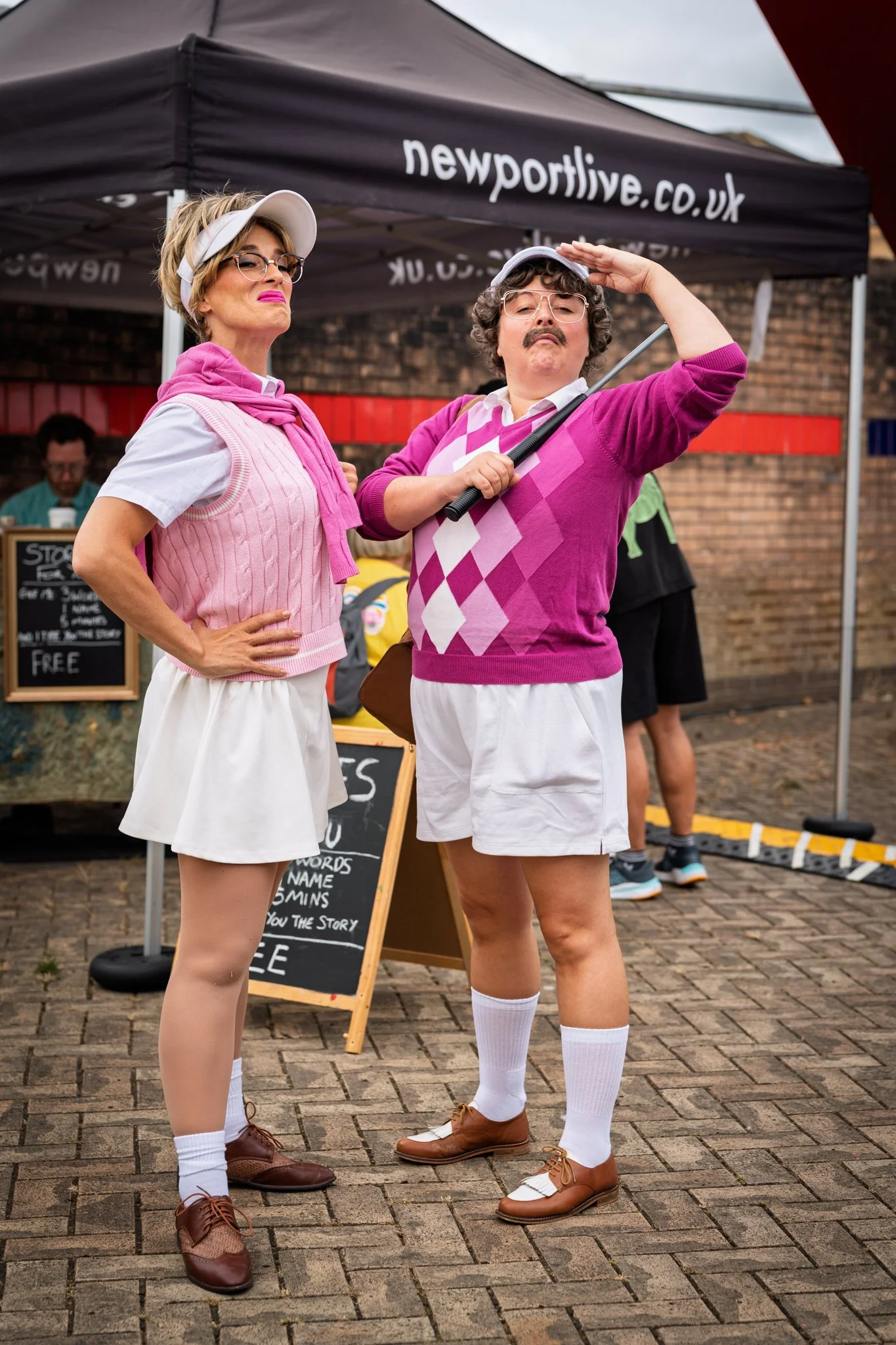Two people dressed as golfers standing outdoors near a black tent with the website 'newportlive.co.uk'. They are wearing retro golf outfits, including white shorts, pink sweaters, knee-high socks, and brown golf shoes. One person is saluting, and the
