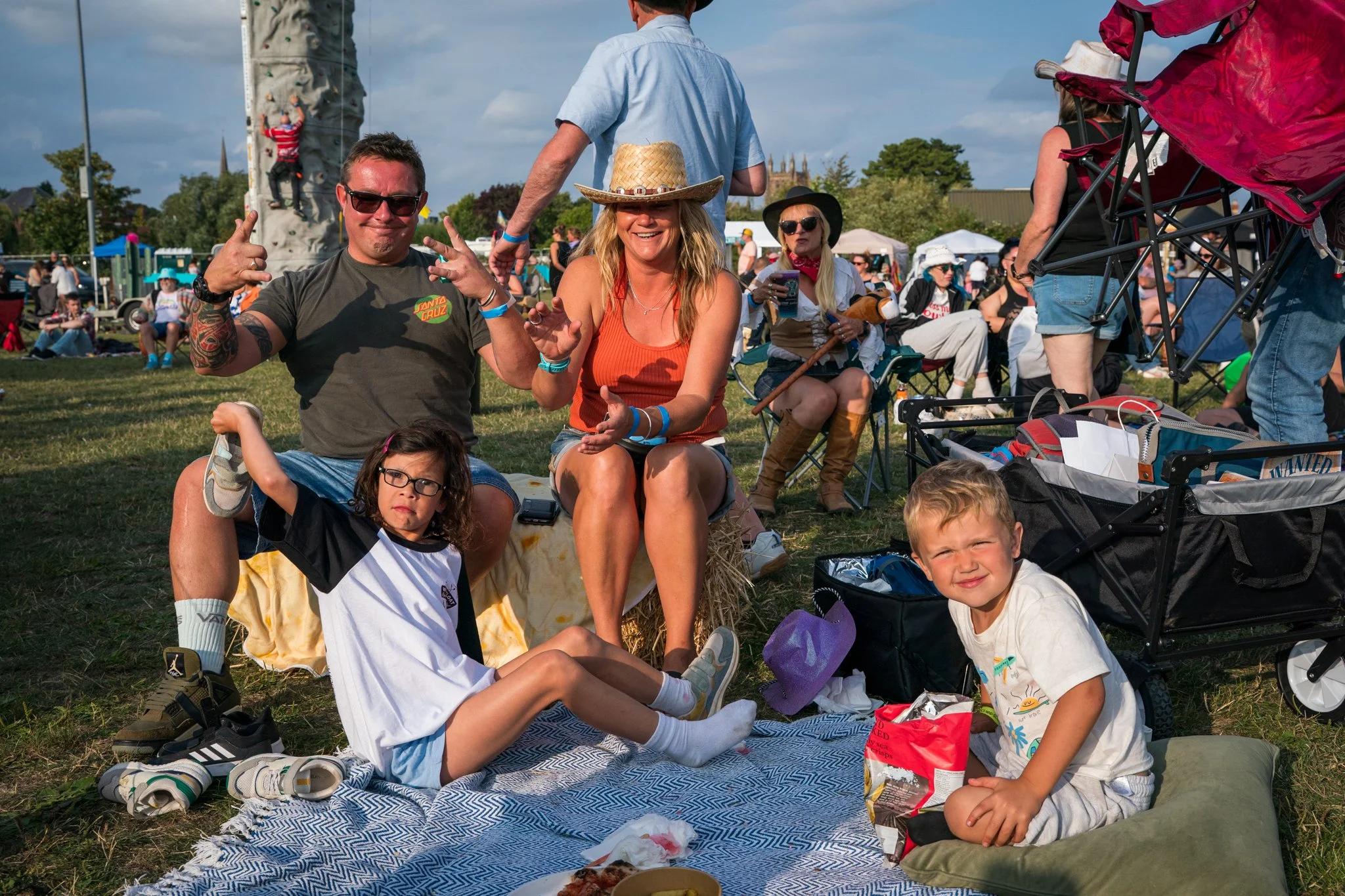Group of people sitting outdoors at a festival or event on a grassy field, with children and adults, some seated on blankets and hay bales, with food and snacks in front.