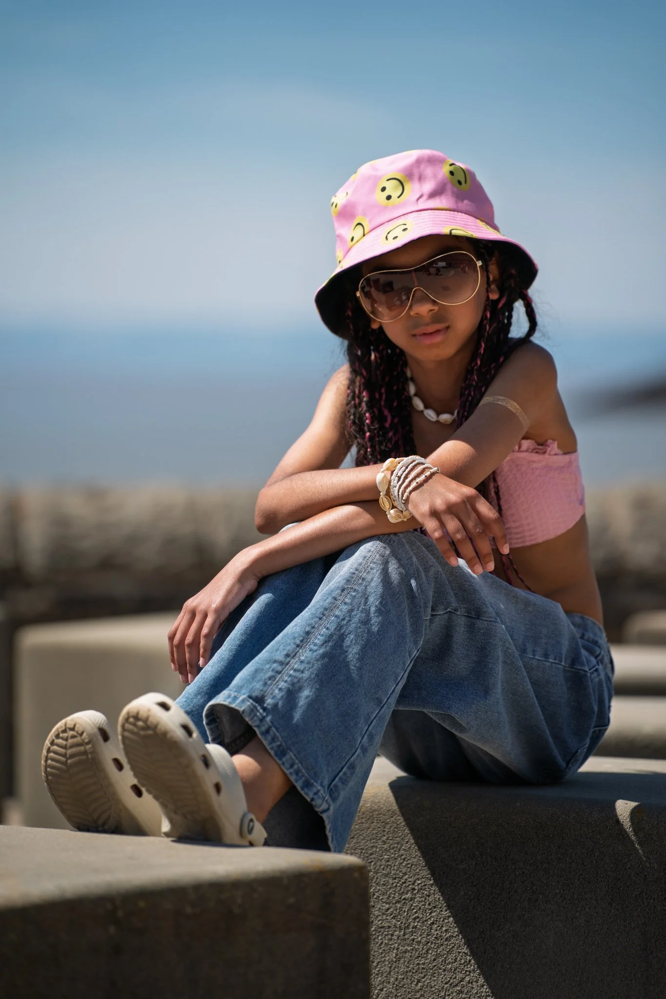 A young girl with dark braided hair, wearing a pink bucket hat with yellow smiley faces, sunglasses, a shell necklace, bracelets, a pink sleeveless top, and blue jeans, sitting on a concrete ledge near the ocean under a clear blue sky.