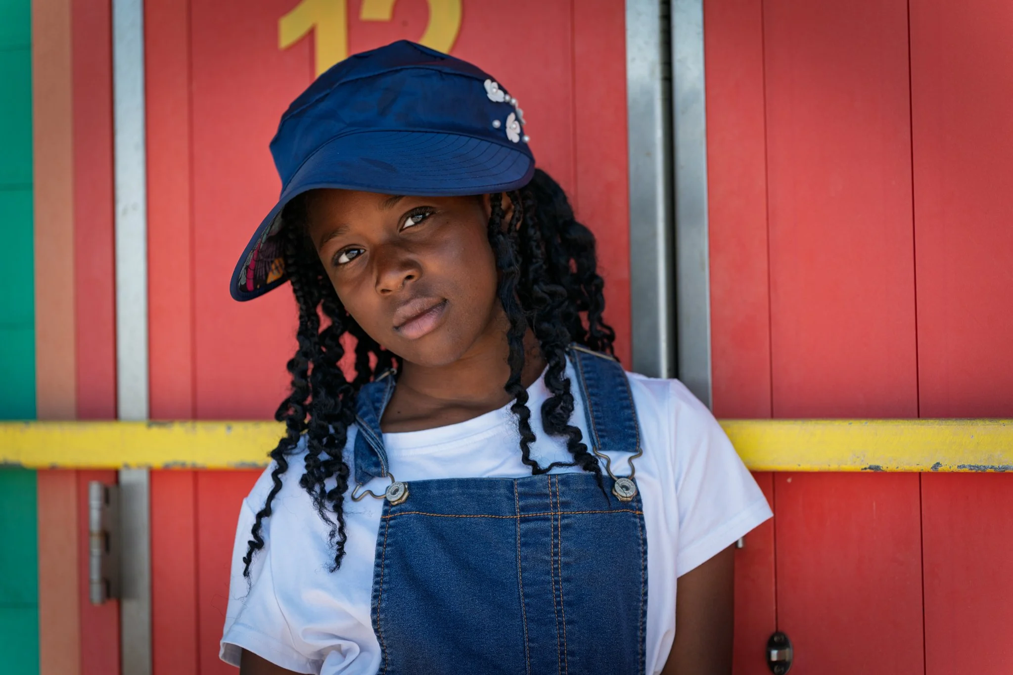 A young girl with dark curly hair wearing a blue cap decorated with white beads and a denim overalls over a white shirt, standing in front of a red wall with colorful panels.