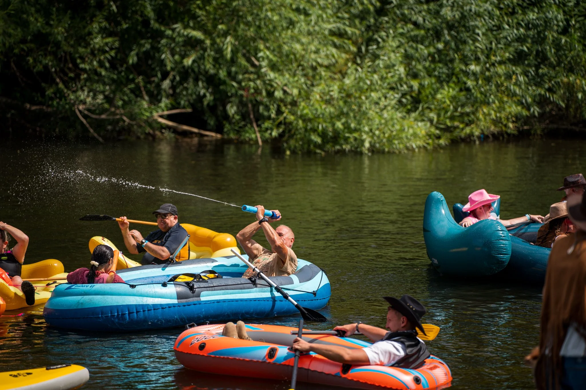 People kayaking and paddleboarding on a river with lush green trees in the background.