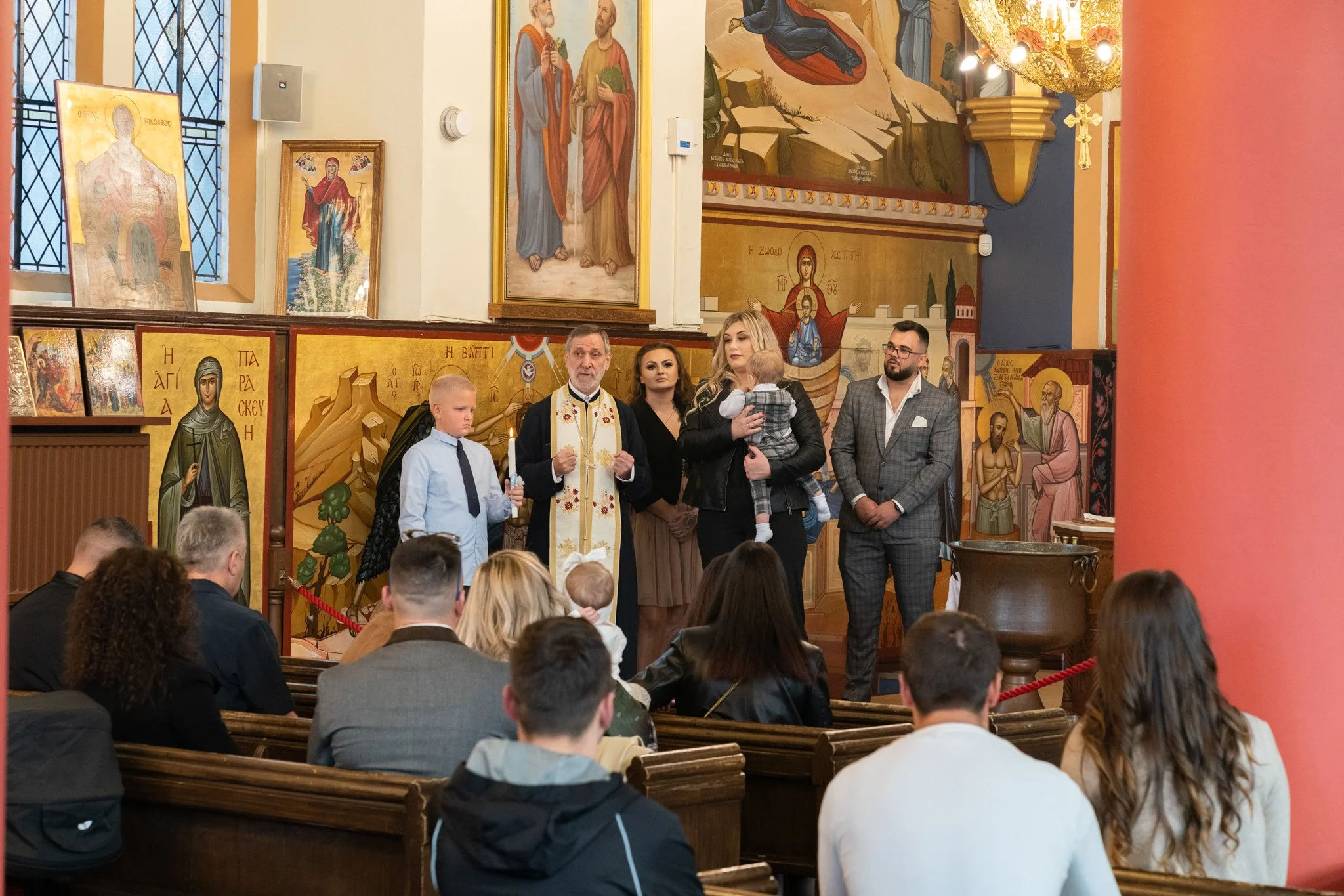 A religious ceremony inside a church with people standing and sitting, including a priest wearing a white stole, several altar icons and religious paintings on the walls, and a woman holding a baby, with a congregation in attendance.