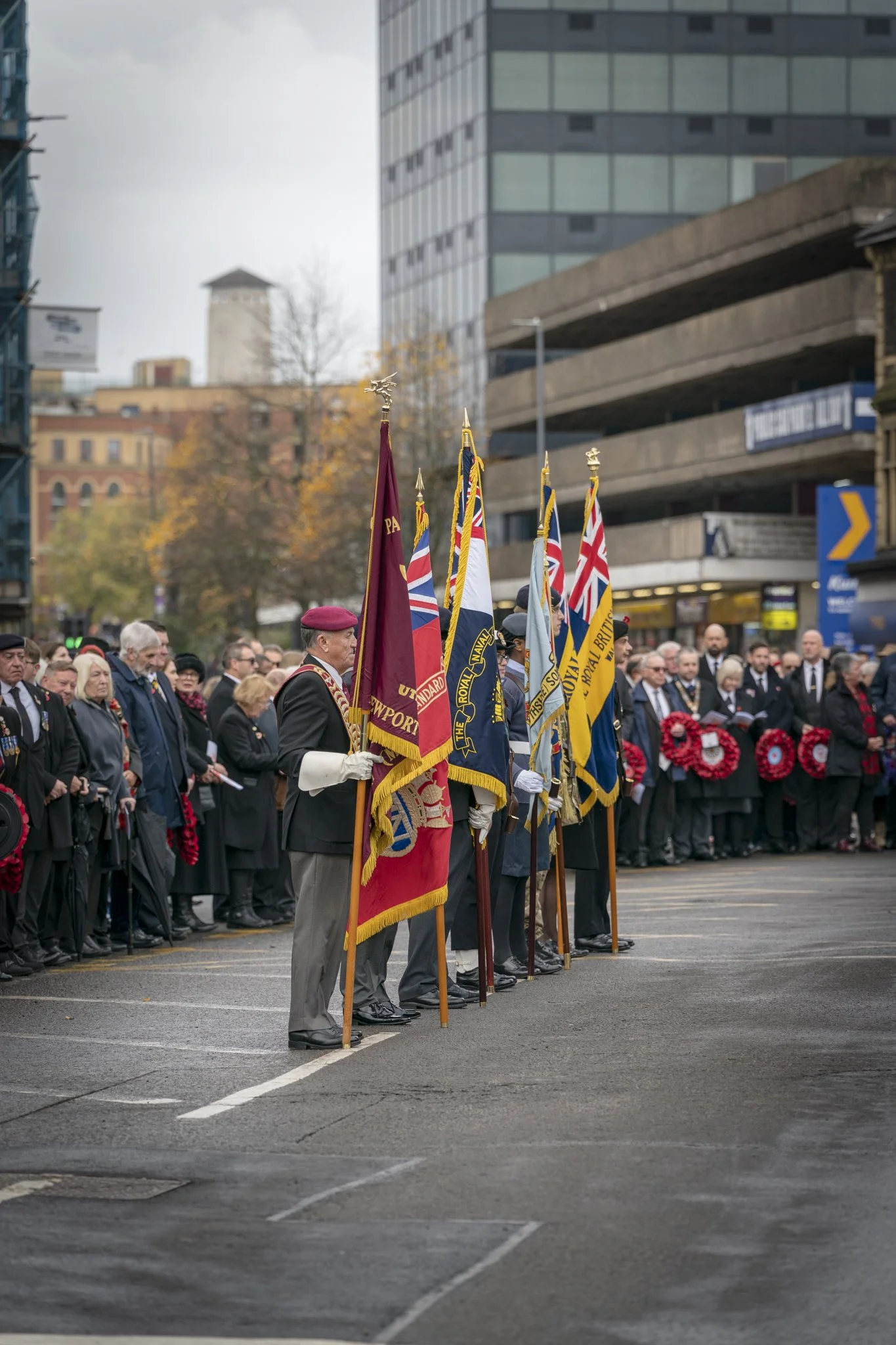 A group of people stand in a line during a ceremony, holding various national flags, with a cityscape of modern and historic buildings in the background.