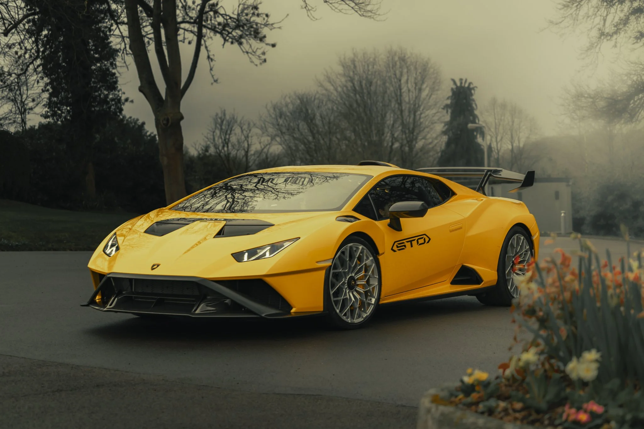 A bright yellow Lamborghini sports car with black accents, parked outdoors on a gray paved road, with trees and overcast sky in the background.