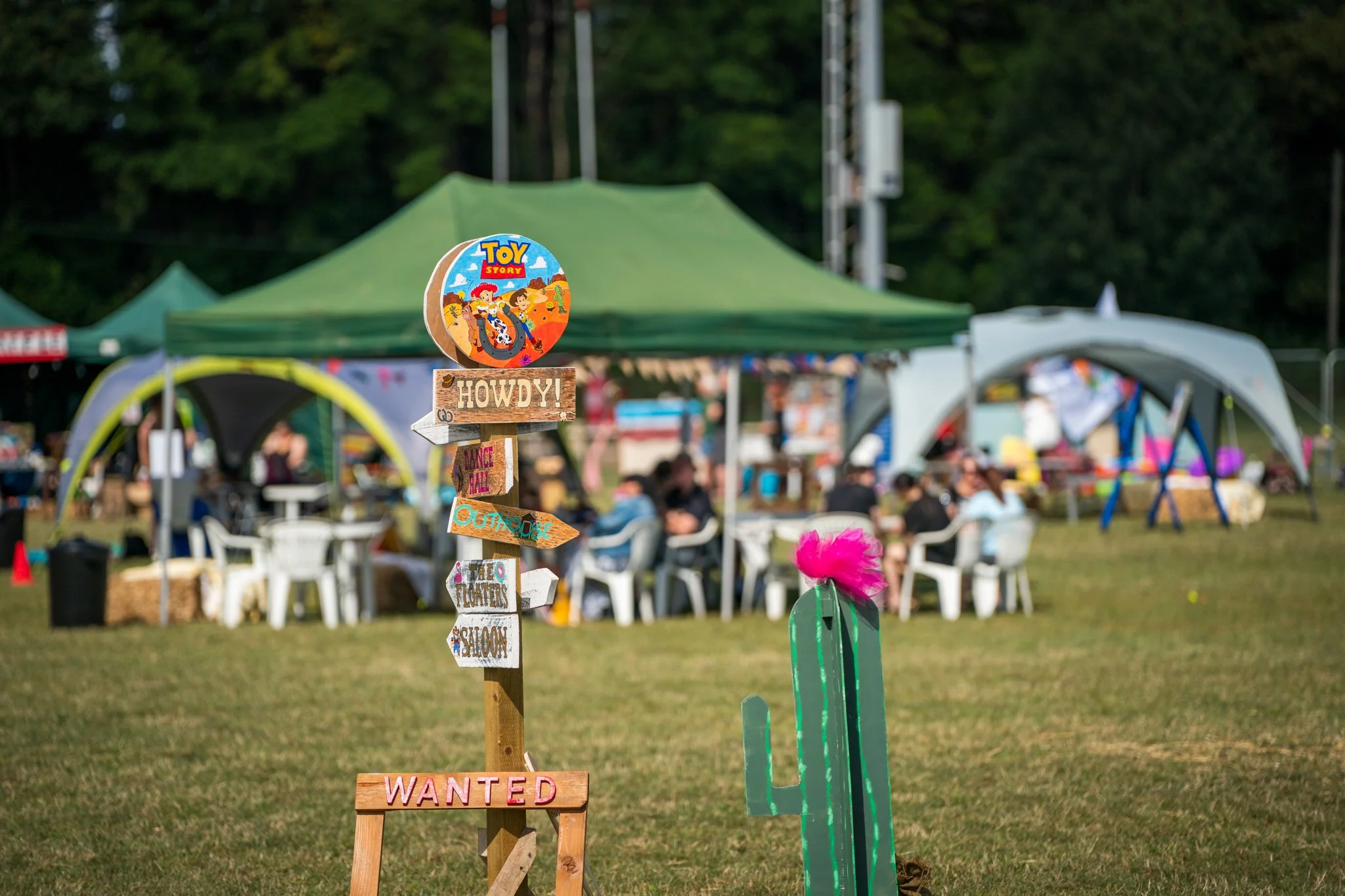 Colorful signs on a wooden post at an outdoor festival or fair, with tents, chairs, and people in the background.