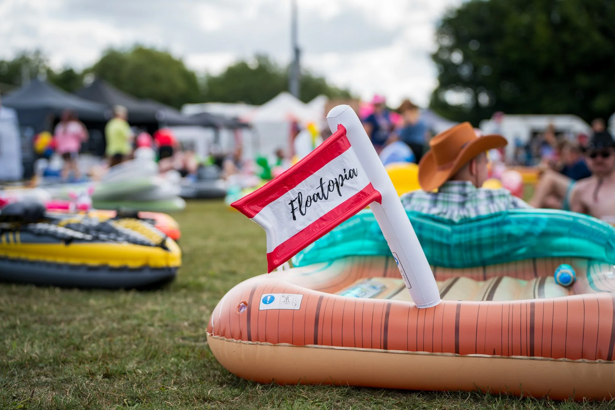 A float in a crowded outdoor event with a small sail flag that says 'Floatopia,' positioned on an inflatable boat or float, with numerous tents and people in the background, indicating a gathering or festival.