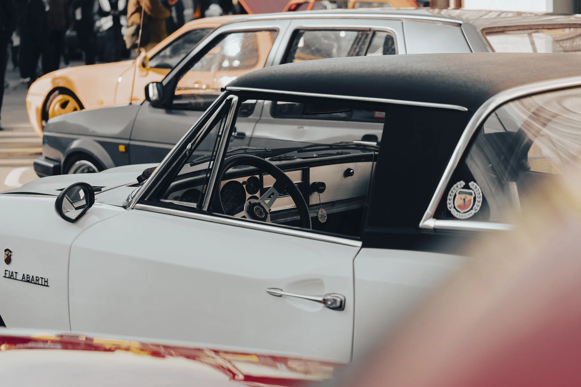 Close-up of a vintage white Fiat Abarth race car with black and orange cars in the background at a car show or event.