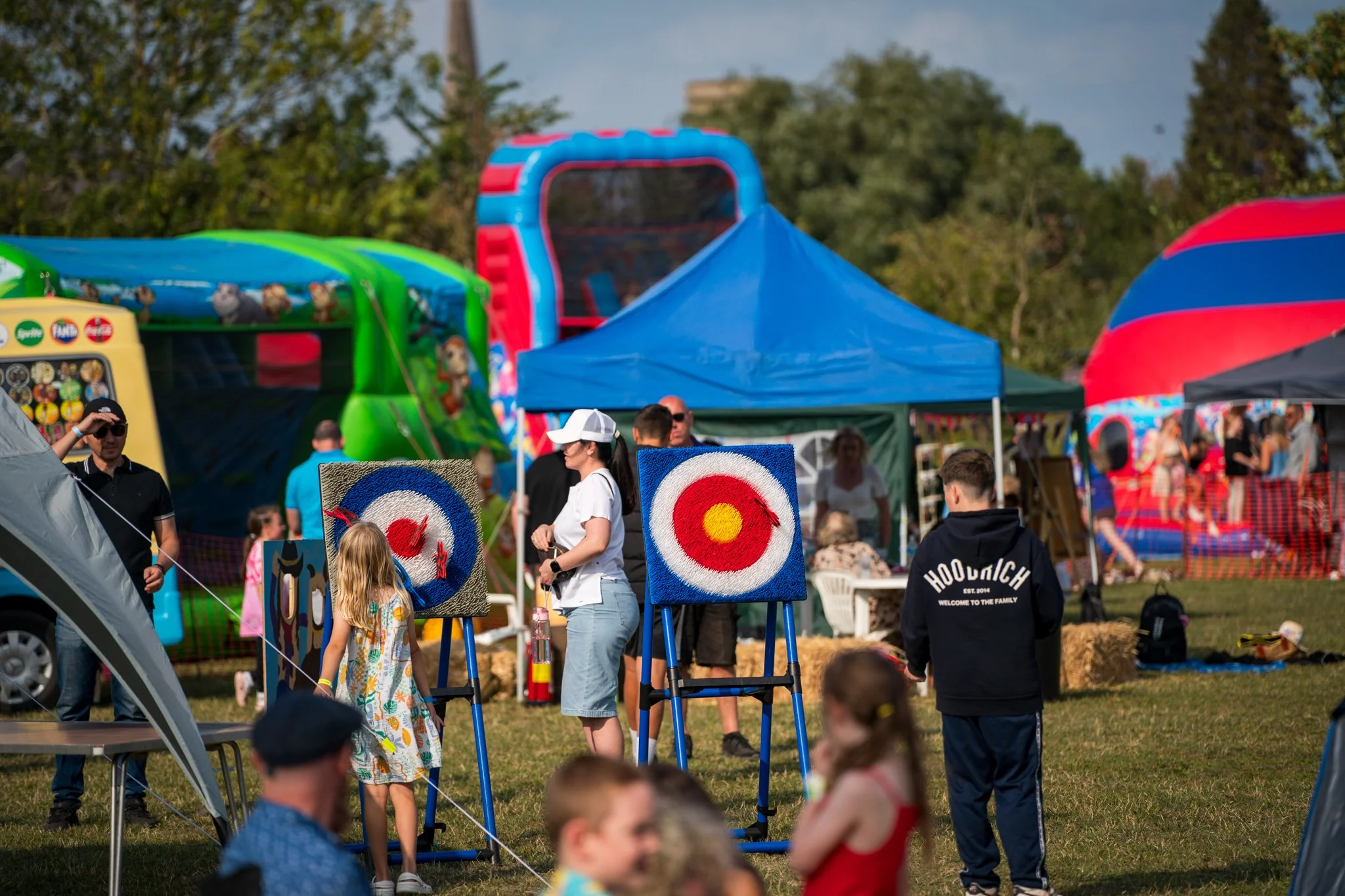 A lively outdoor fair with people participating in a target throwing game. The scene features colorful tents and inflatable rides in the background, with trees and a blue sky overhead.