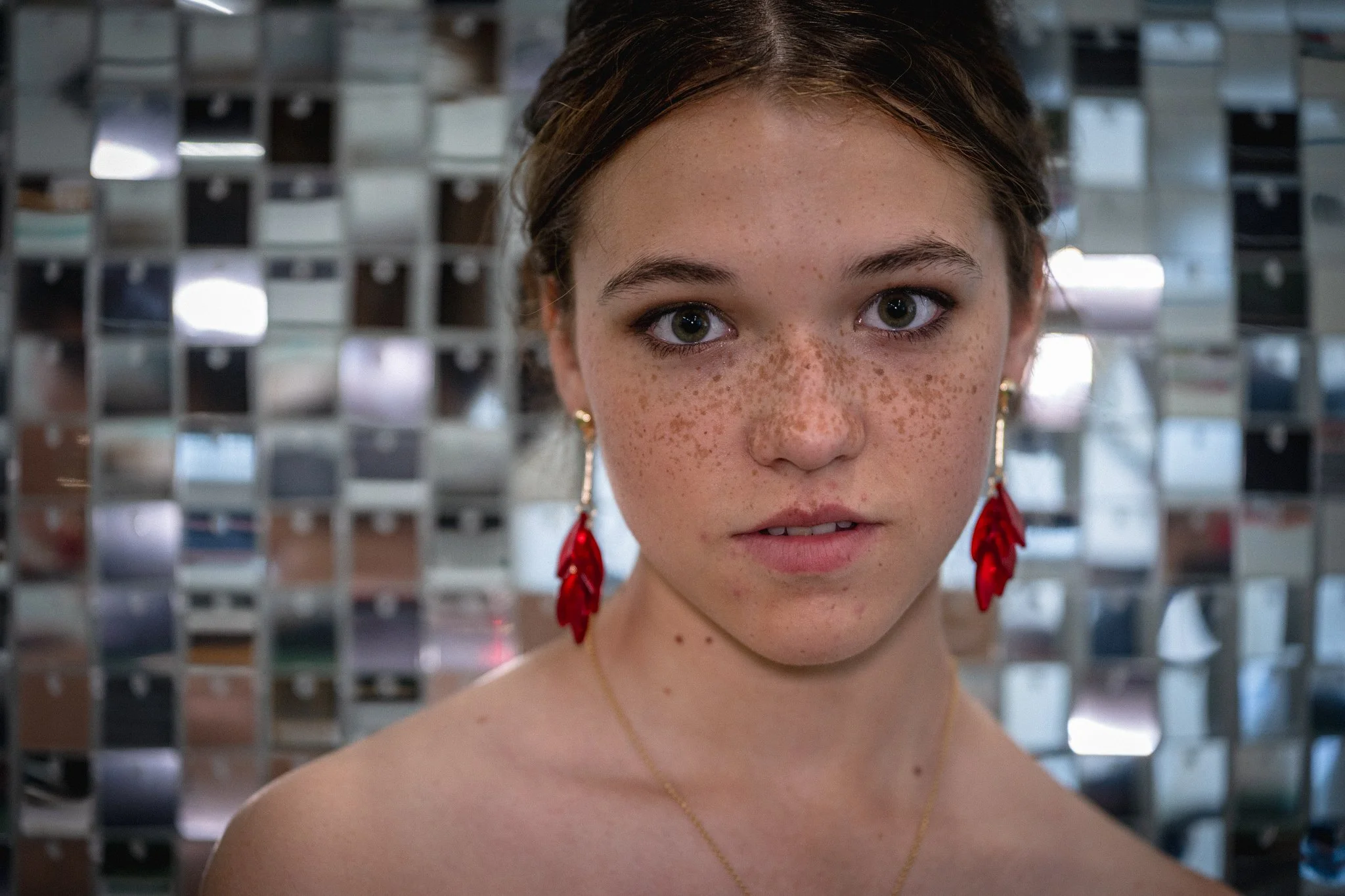 A young woman with freckles and green eyes looking directly at the camera, partially framed by a metallic grid background. She has dark hair pulled back and is wearing red earrings and a necklace.