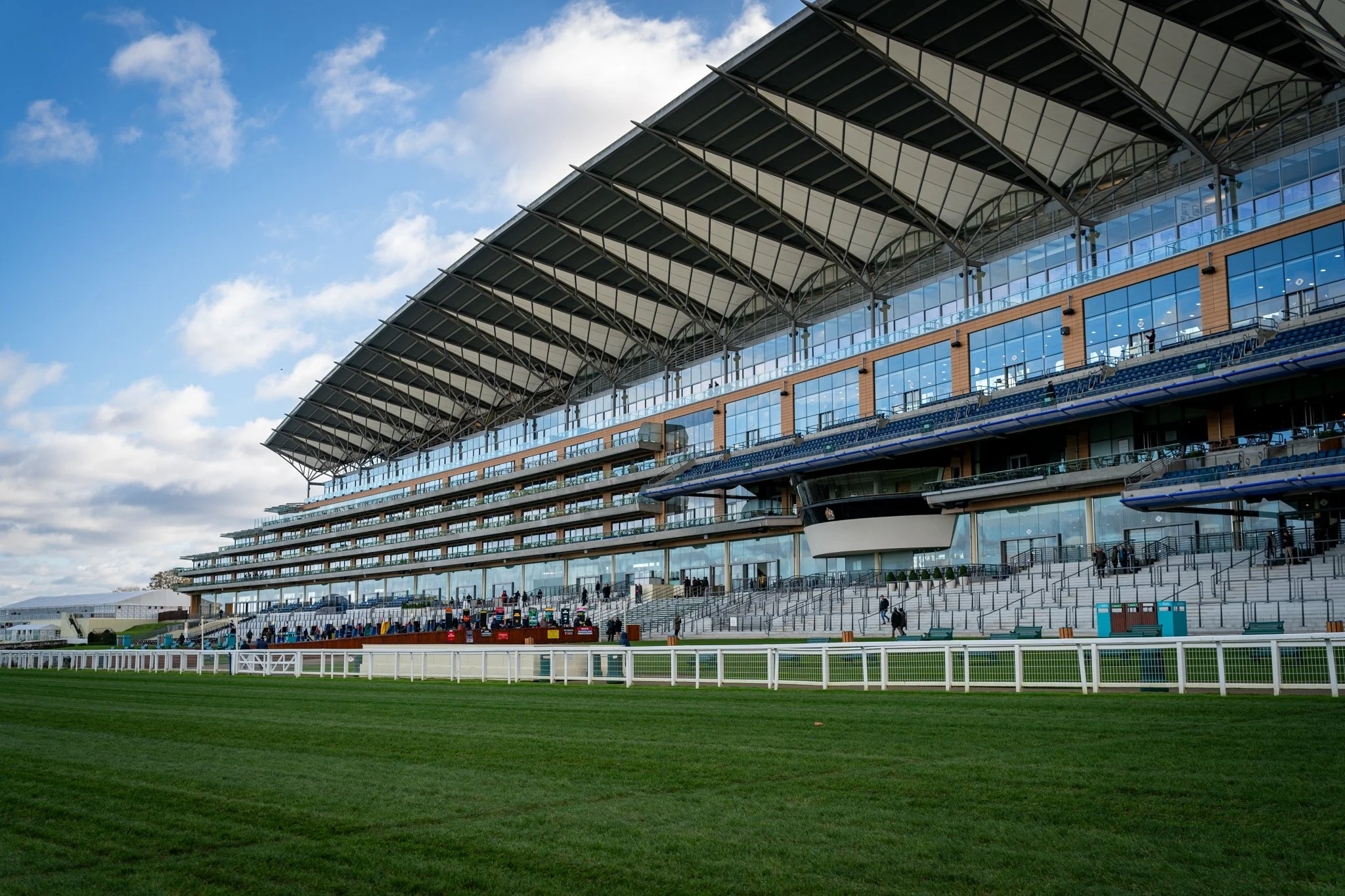 View of a modern horse racing grandstand with multiple tiers of seating, large glass windows, and a distinctive angled roof, set against a partly cloudy sky and overlooking a green racecourse.