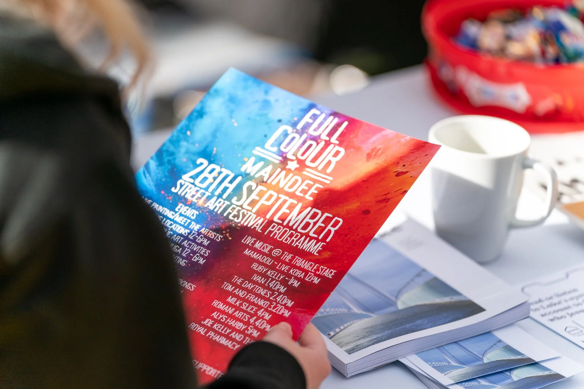 A person holding a colorful event flyer, with a background of a table with a white mug and a red bowl filled with small objects or candies.