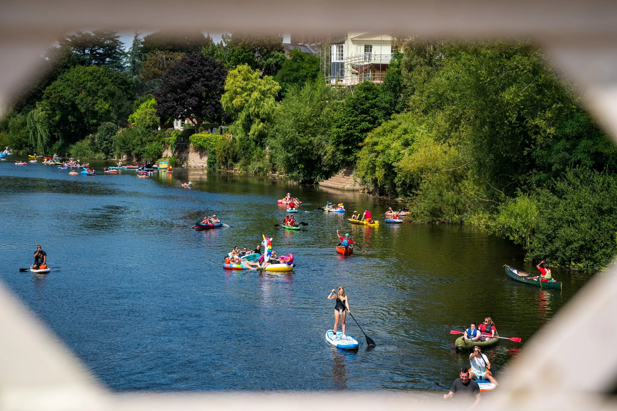 People paddleboarding and kayaking on a river surrounded by lush green trees and houses, viewed through an opening.