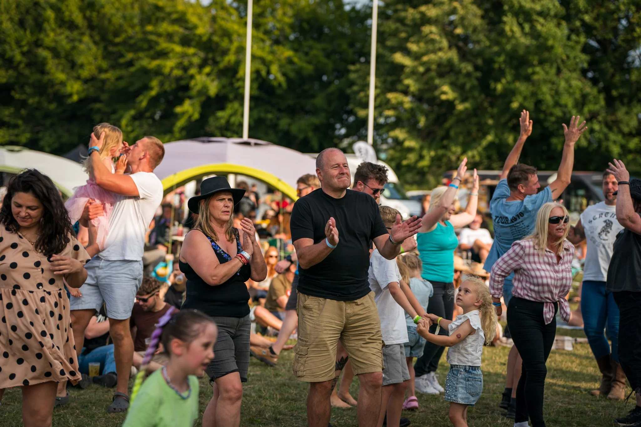 People enjoying an outdoor event, some dancing and watching the performances with trees and tents in the background.