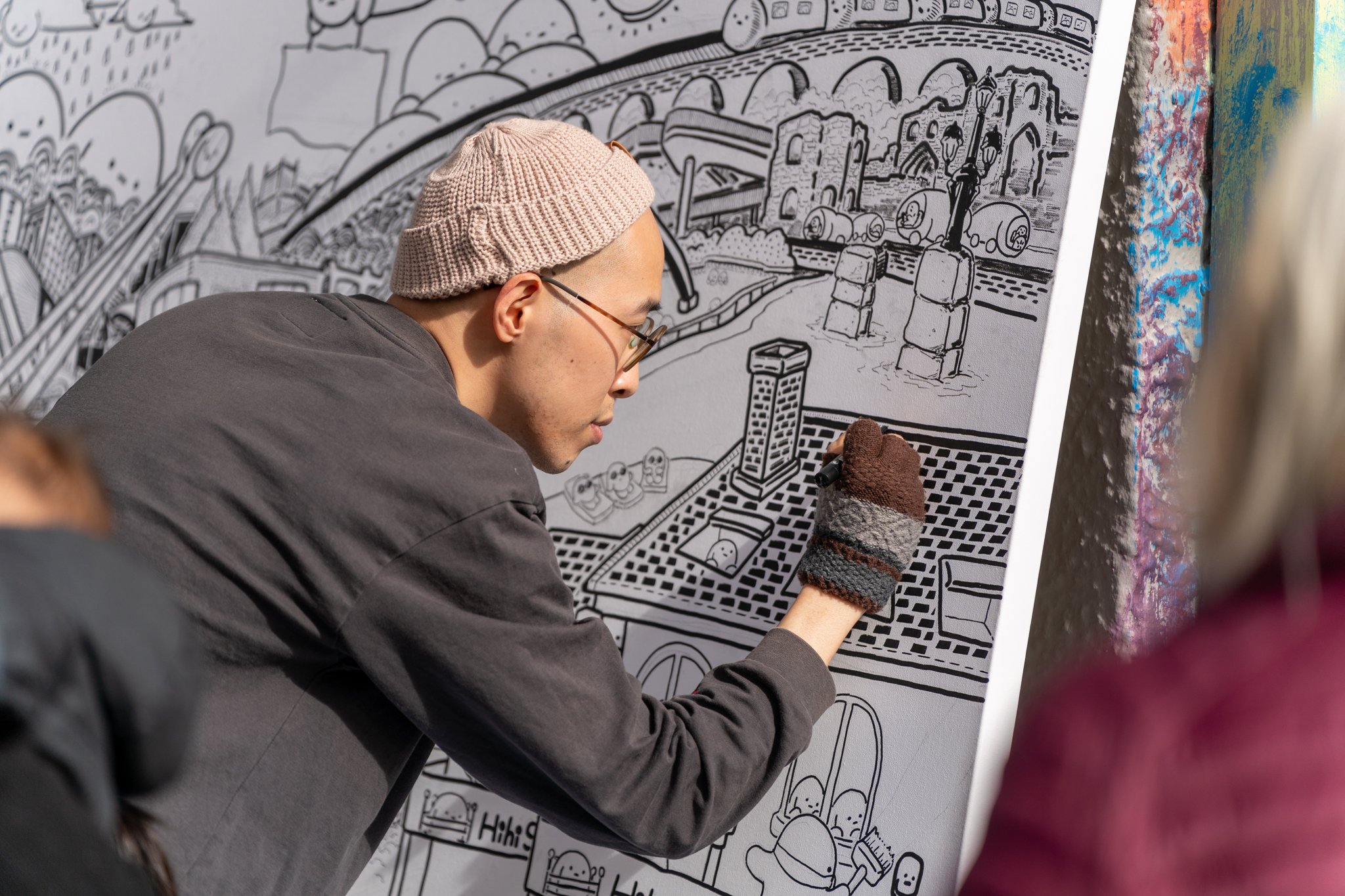 A person wearing glasses, a pink beanie, and a dark jacket is drawing on a large black and white mural with a marker. The mural features a cityscape with bridges, buildings, trains, and various cartoon characters.