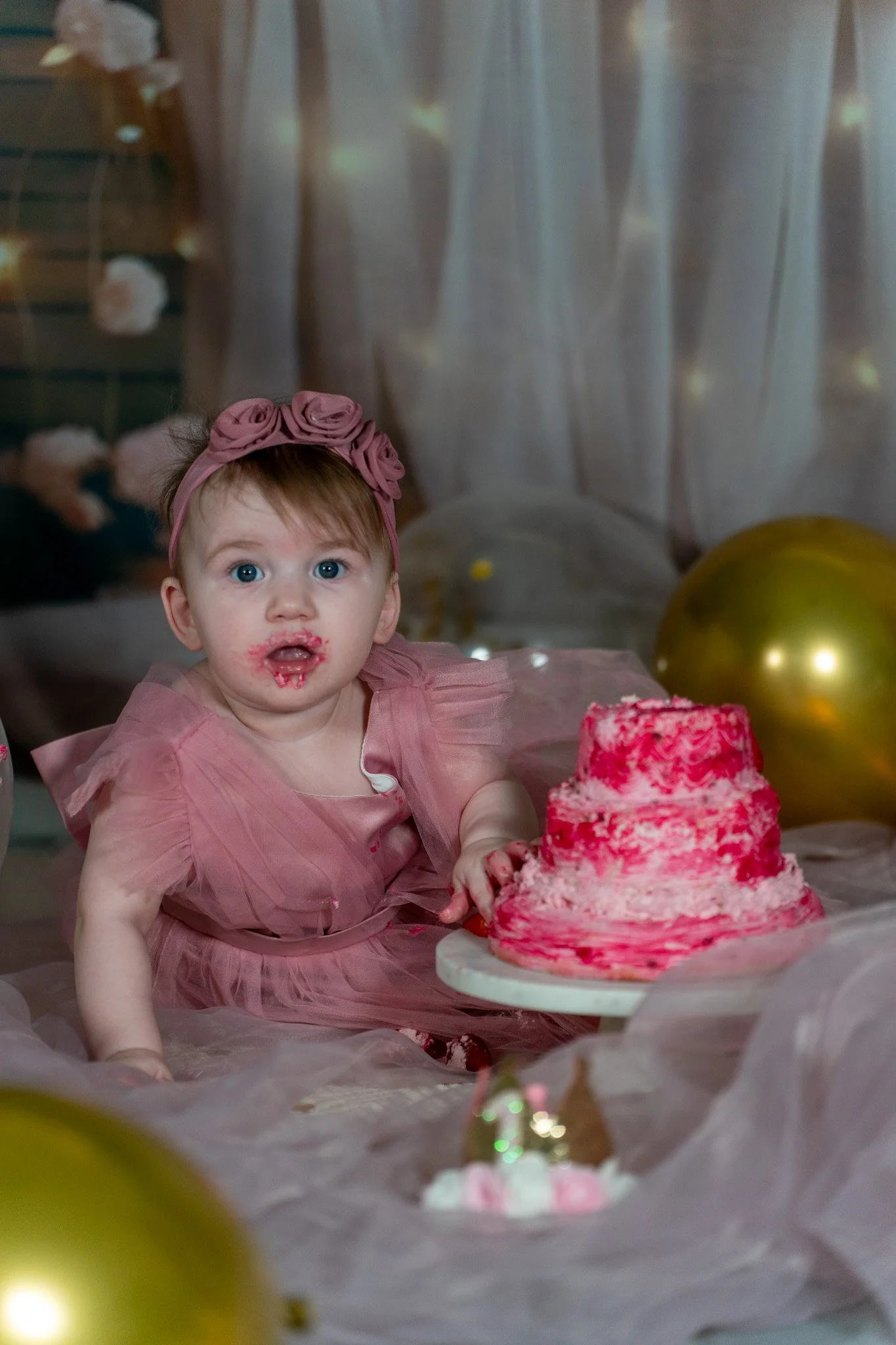 A young girl with blue eyes and a pink headband with roses, wearing a pink dress, sits in front of a pink birthday cake with red streaks. She has some frosting on her face and hand, and in front of her are balloons and birthday decorations, indicatin