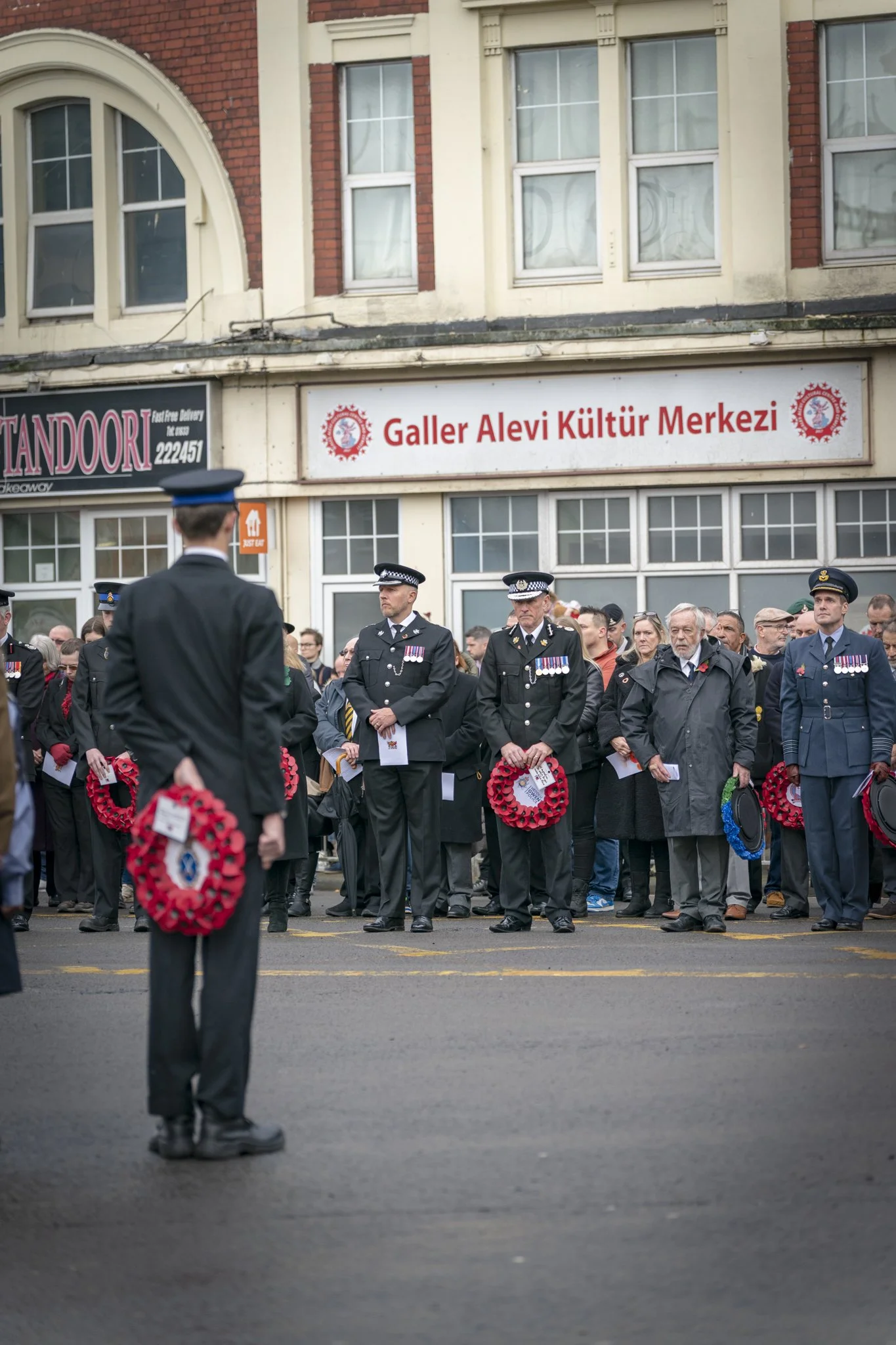 A crowd of people, including several uniformed officers, gathered for a memorial or remembrance event in front of a building with signs labeled "Galler Alevi Kultur Merkezi" and other shops. Many people are holding or wearing wreaths or flowers, indi