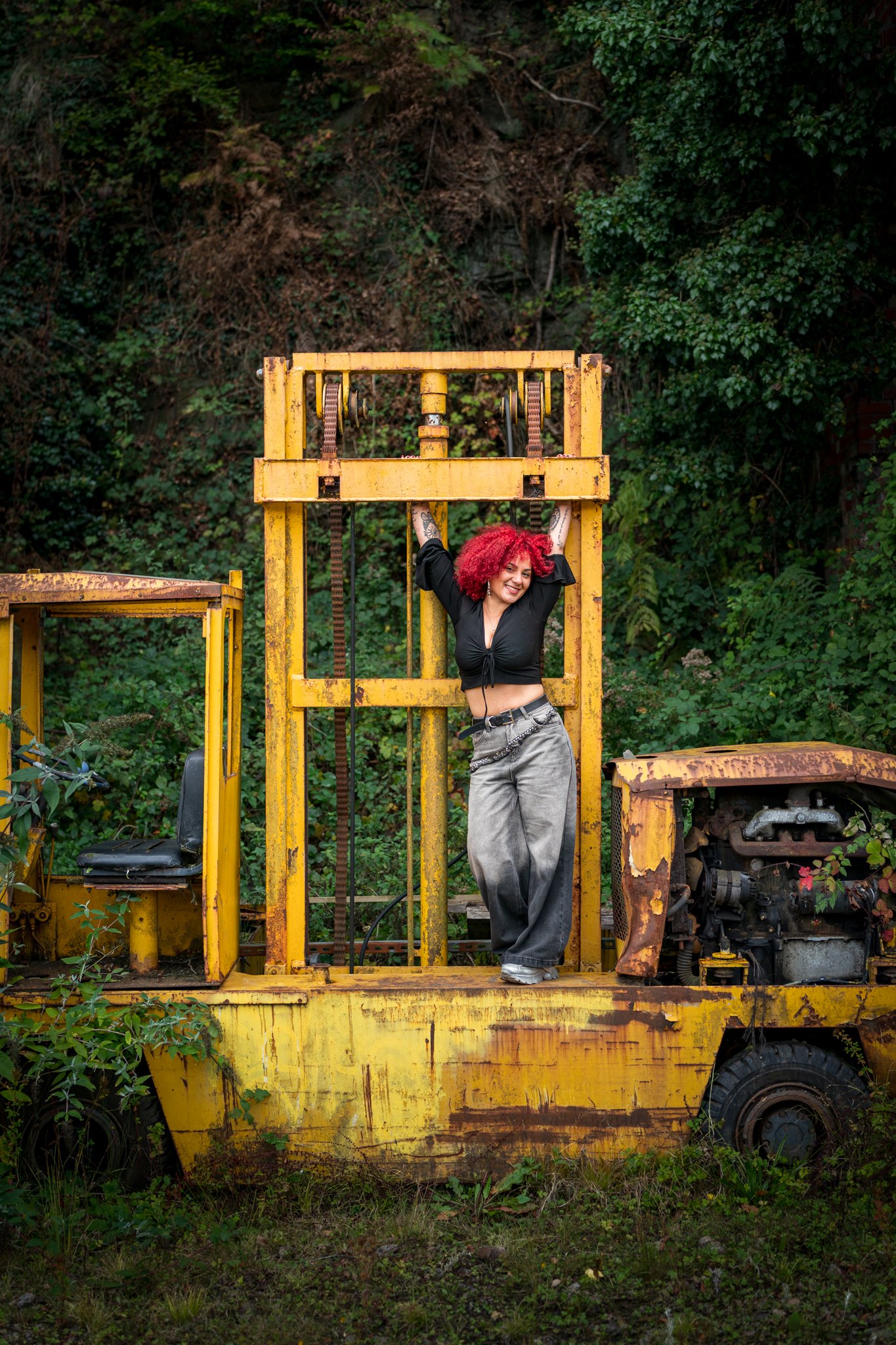 A woman with curly red hair smiles and poses with her arms raised behind a yellow, rusty construction or industrial machine or vehicle in an outdoor setting with green foliage in the background.