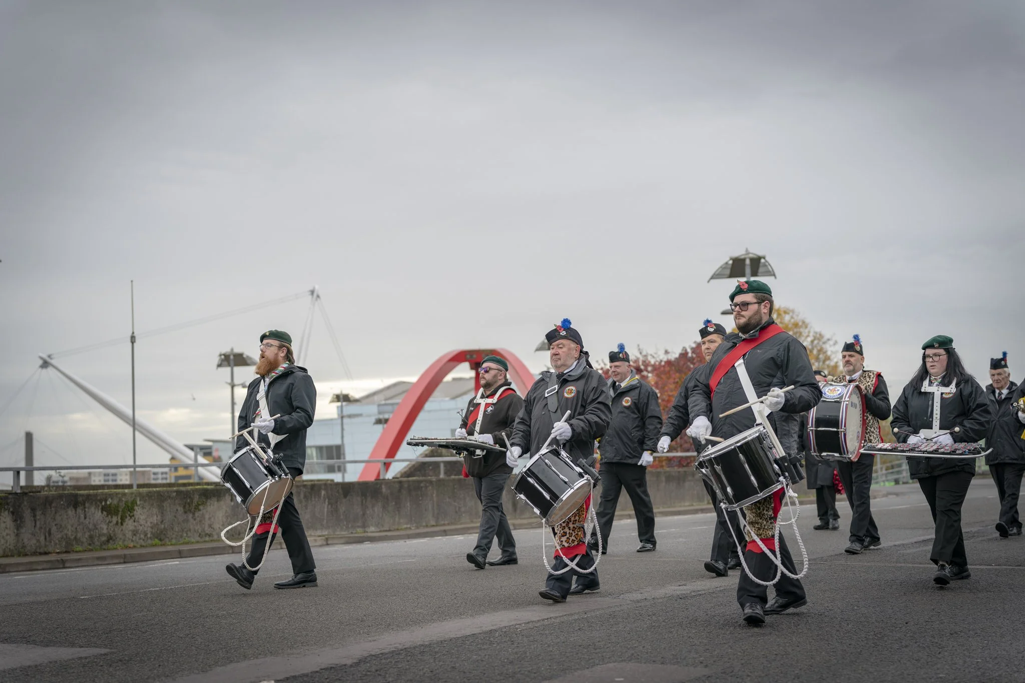 A group of musicians marching outdoors, playing drums and percussion instruments, dressed in coordinated dark uniforms with red and white accents, and wearing berets with decorative pins, under a cloudy sky.