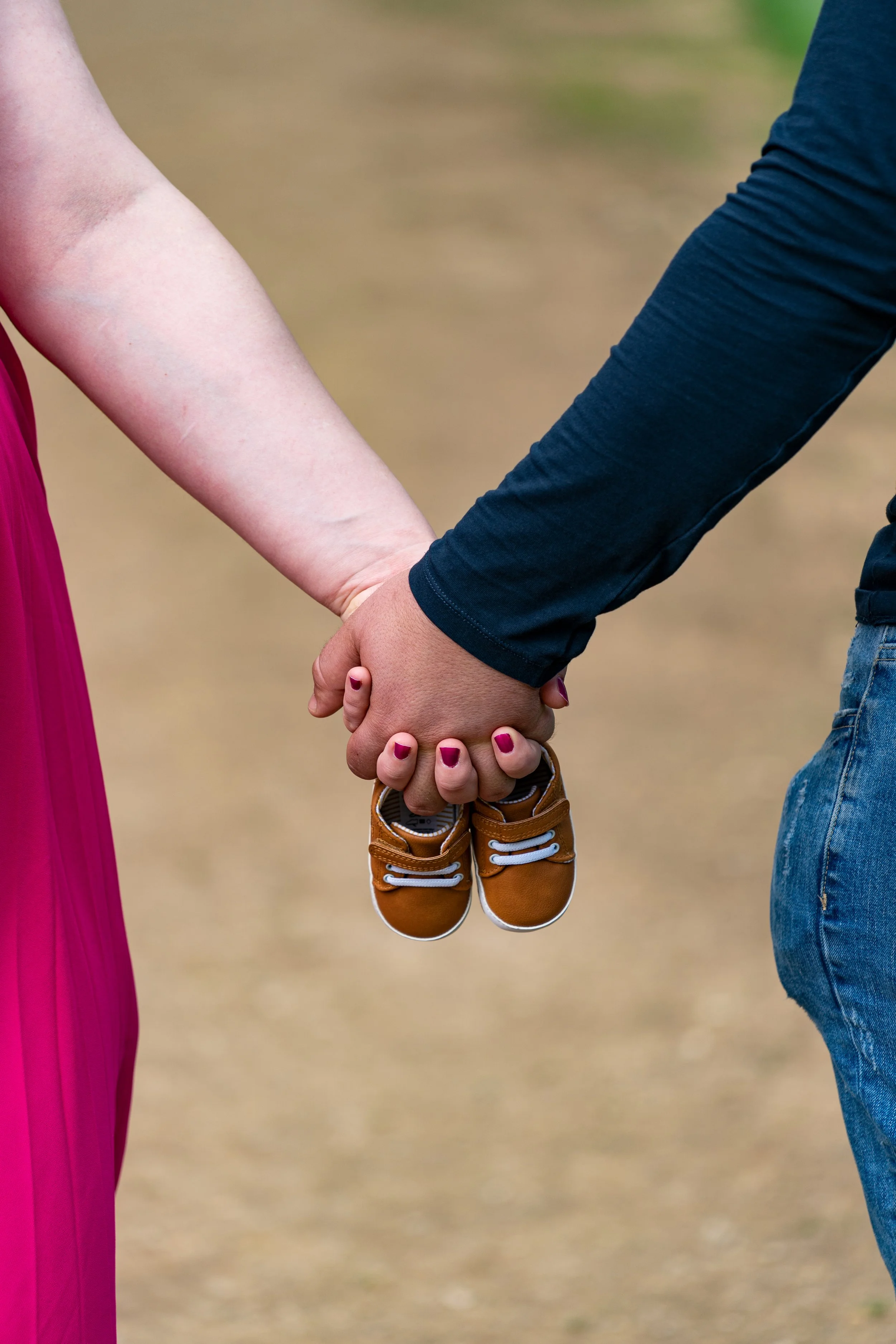 A person wearing a dark blue long-sleeve shirt and jeans holding the hand of a woman in a pink dress. The woman is also holding a pair of small brown shoes.