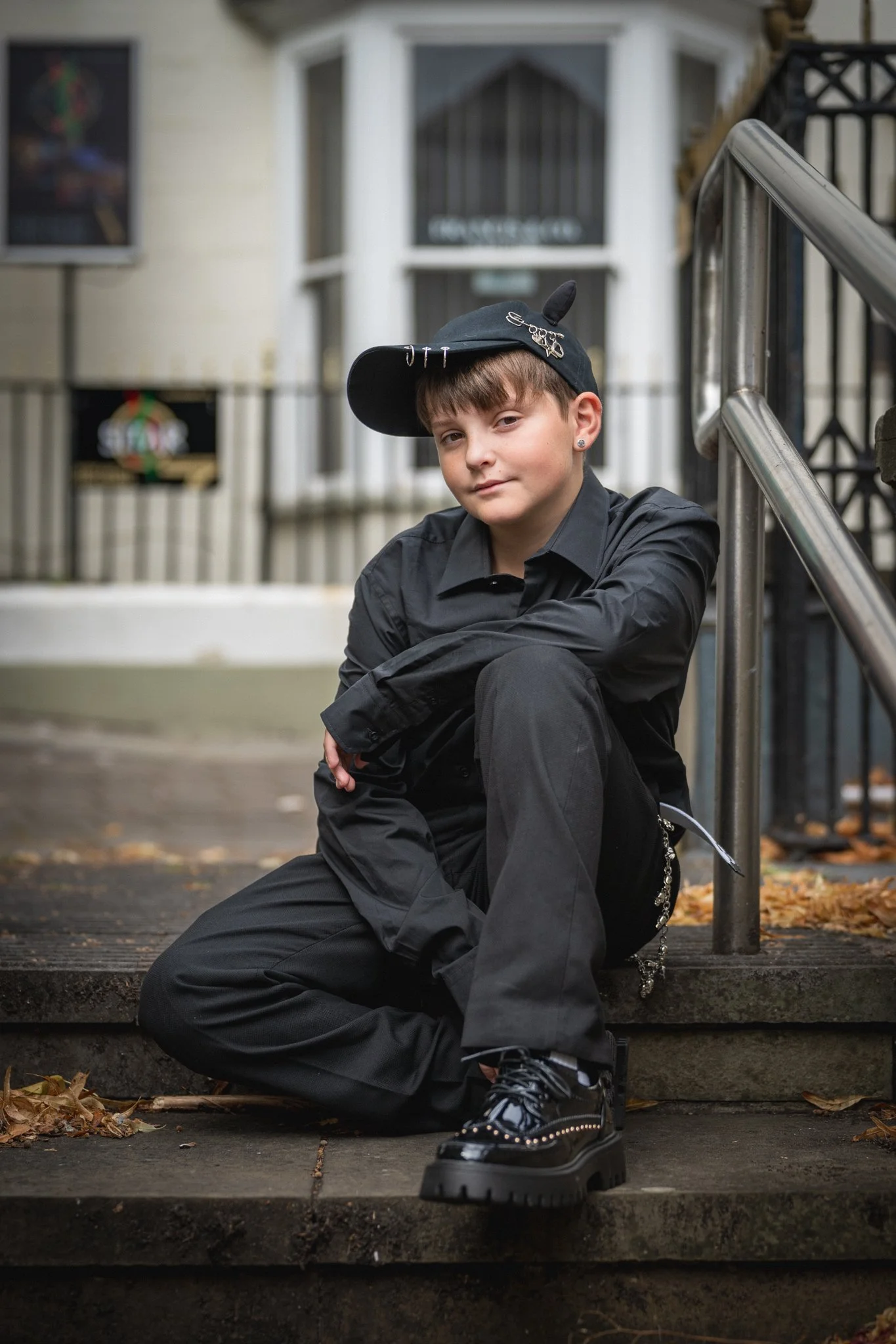 A young person with brown hair and earrings sitting on outdoor stairs with a modern black outfit, black boots, and a baseball cap decorated with pins and chain embellishments.