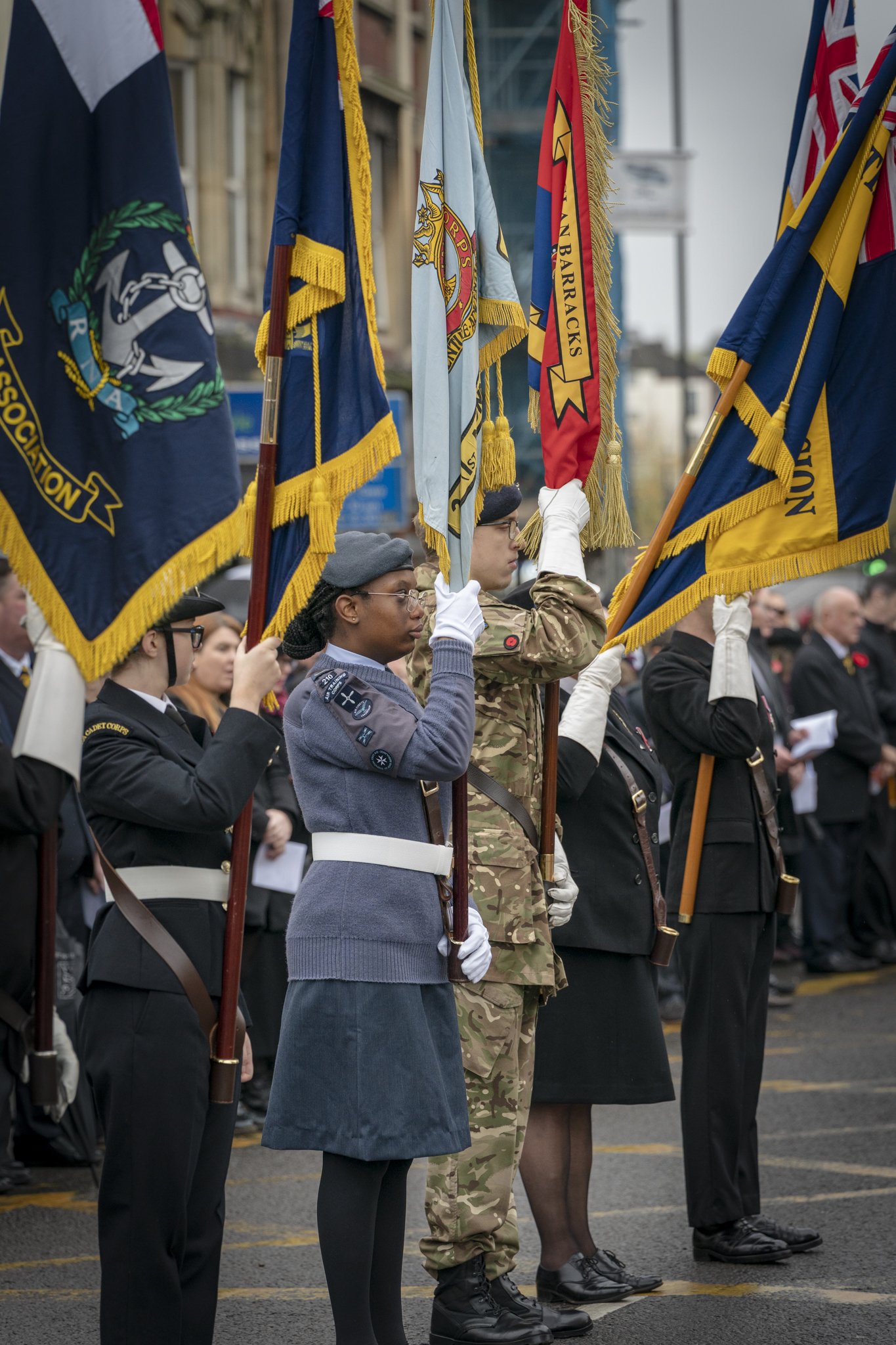 People in military and service uniforms standing in a line holding flags during a parade or ceremony, with a crowd in the background.