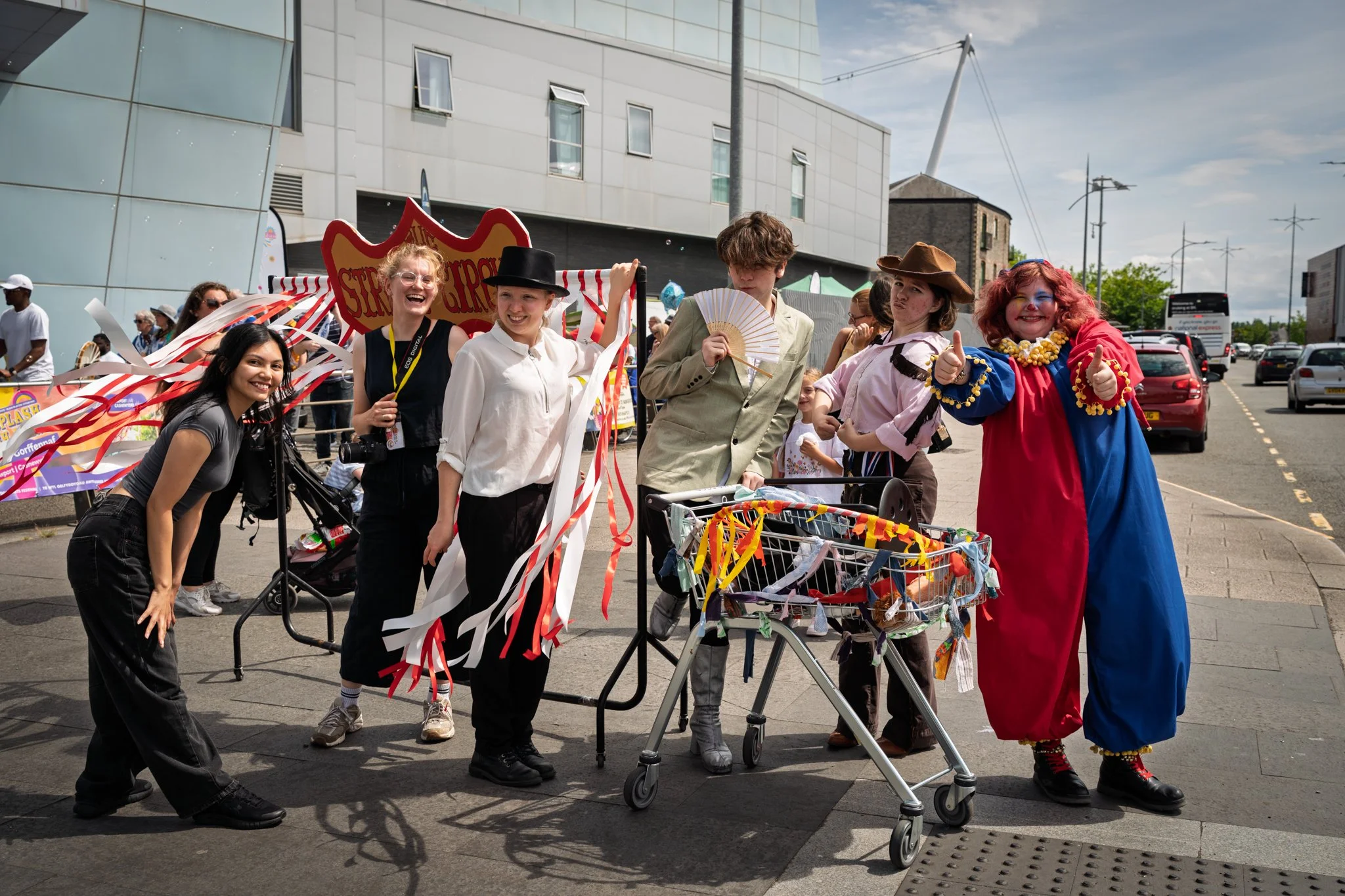 A group of people dressed in colorful costumes participating in a parade or festival outdoors, with some holding items like fans and a shopping cart decorated with ribbons, smiling and giving thumbs up.
