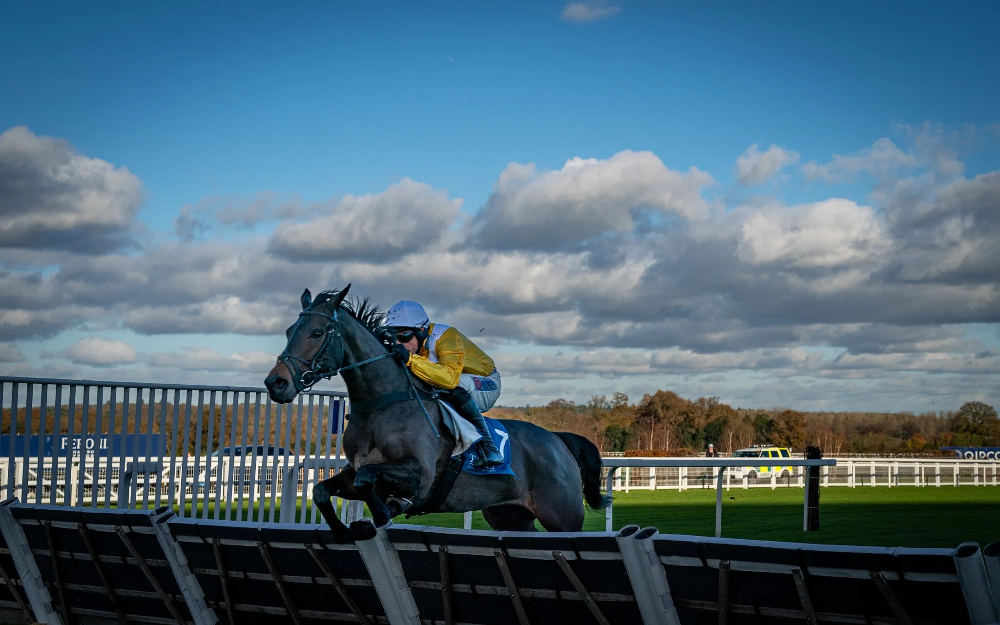 A jockey in a yellow and white uniform and helmet riding a dark gray horse on a racetrack under a partly cloudy sky.